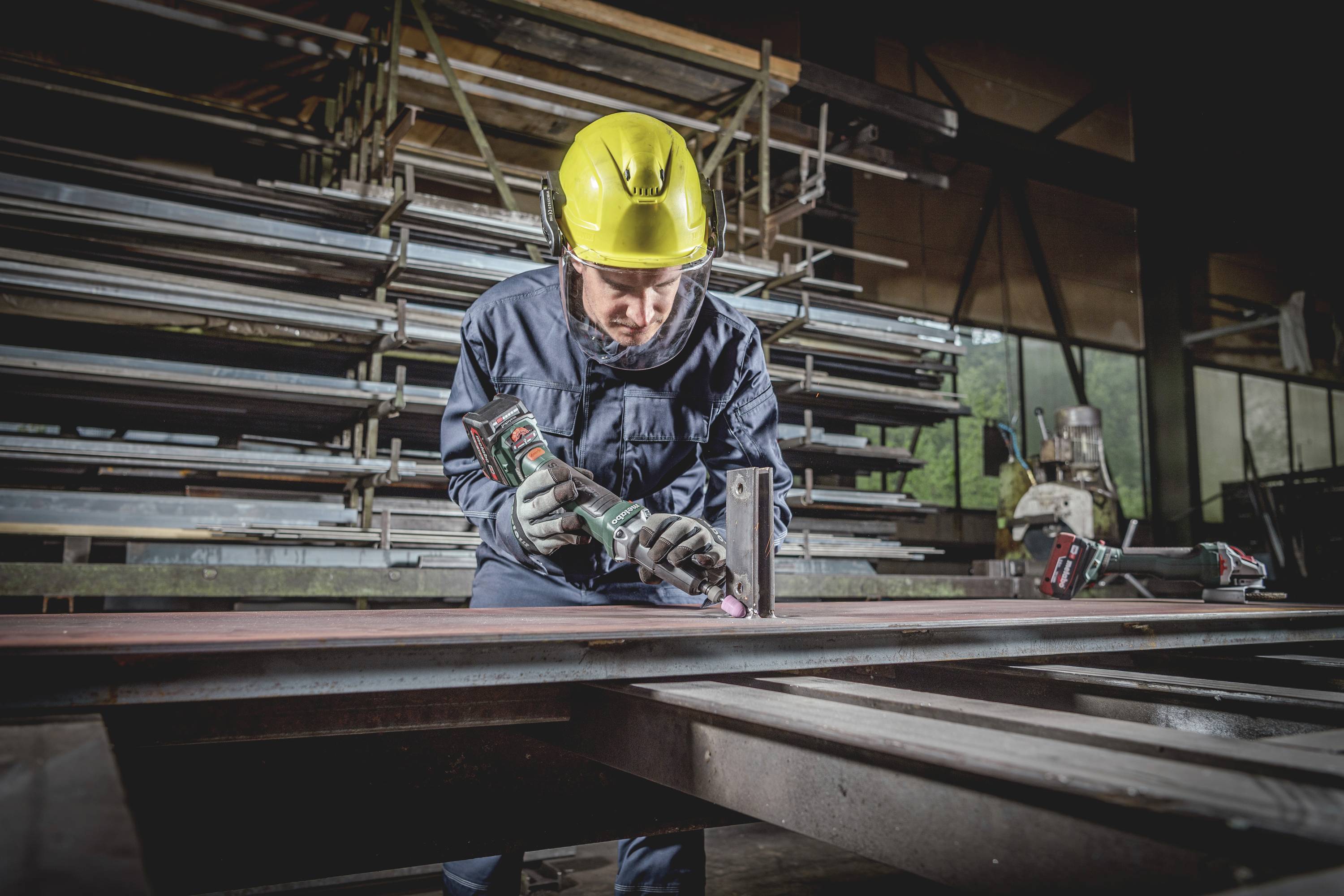 Ein Bauarbeiter in Schutzkleidung und gelbem Helm bohrt mit einer Maschine in einem Metallwerk. Im Hintergrund sind Metallgerüste.