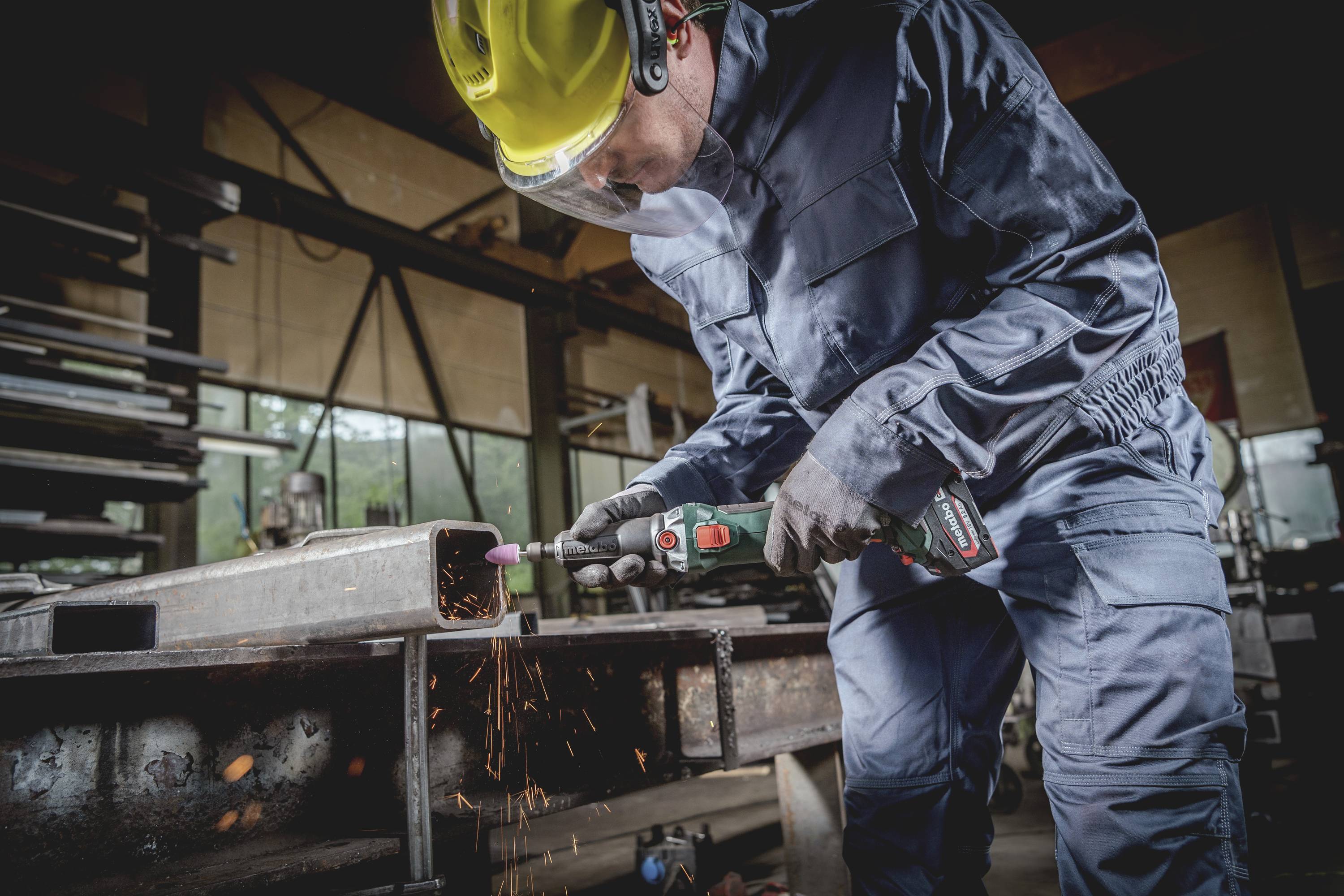 Eine Person in blauer Arbeitskleidung und Helm schleift mit einem Elektrowerkzeug ein Metallstück in einer Werkstatt. Funken fliegen.
