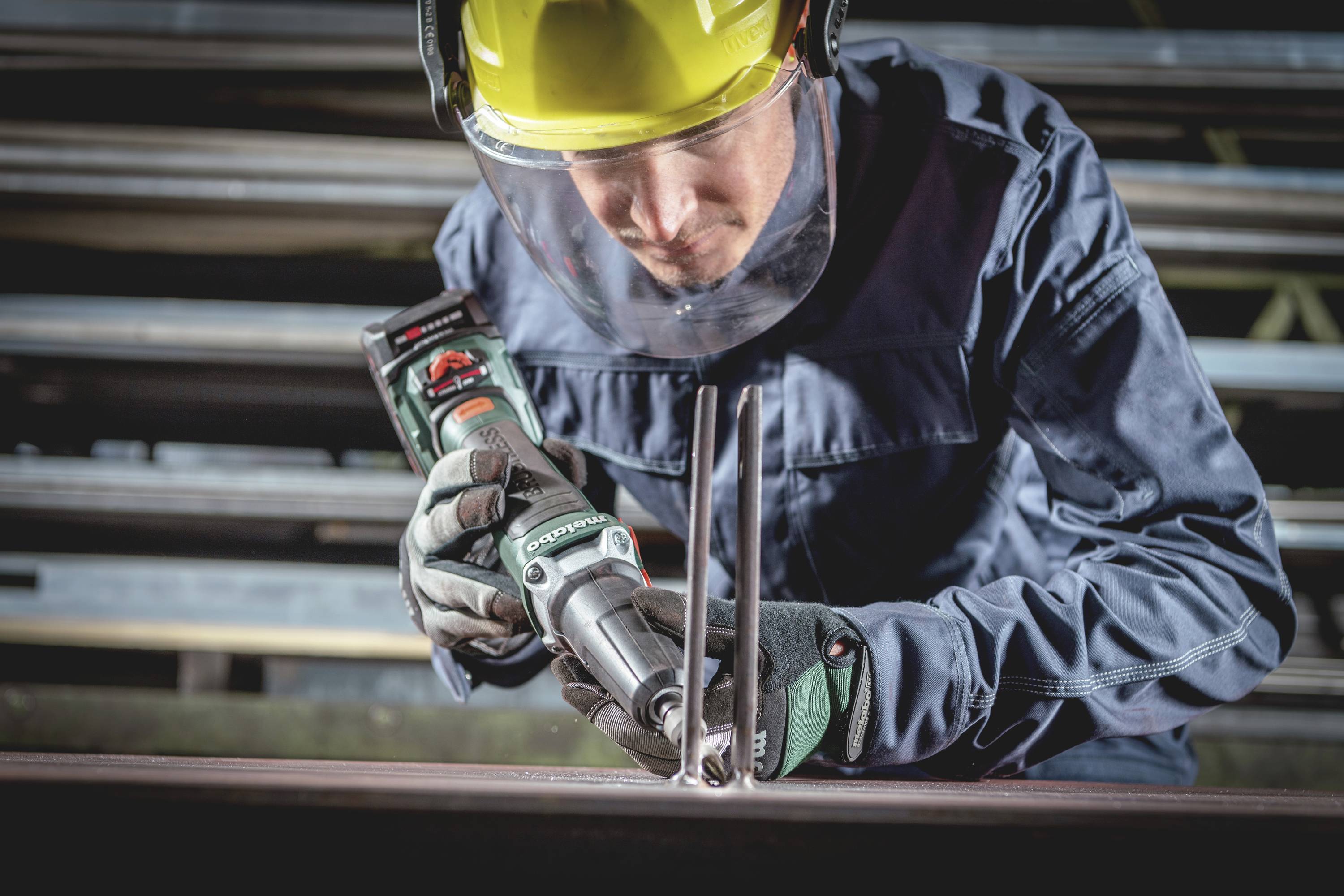 Ein Arbeiter trägt Schutzkleidung, benutzt ein Elektrowerkzeug zum Schleifen von Metallstäben in einer Werkstattumgebung.