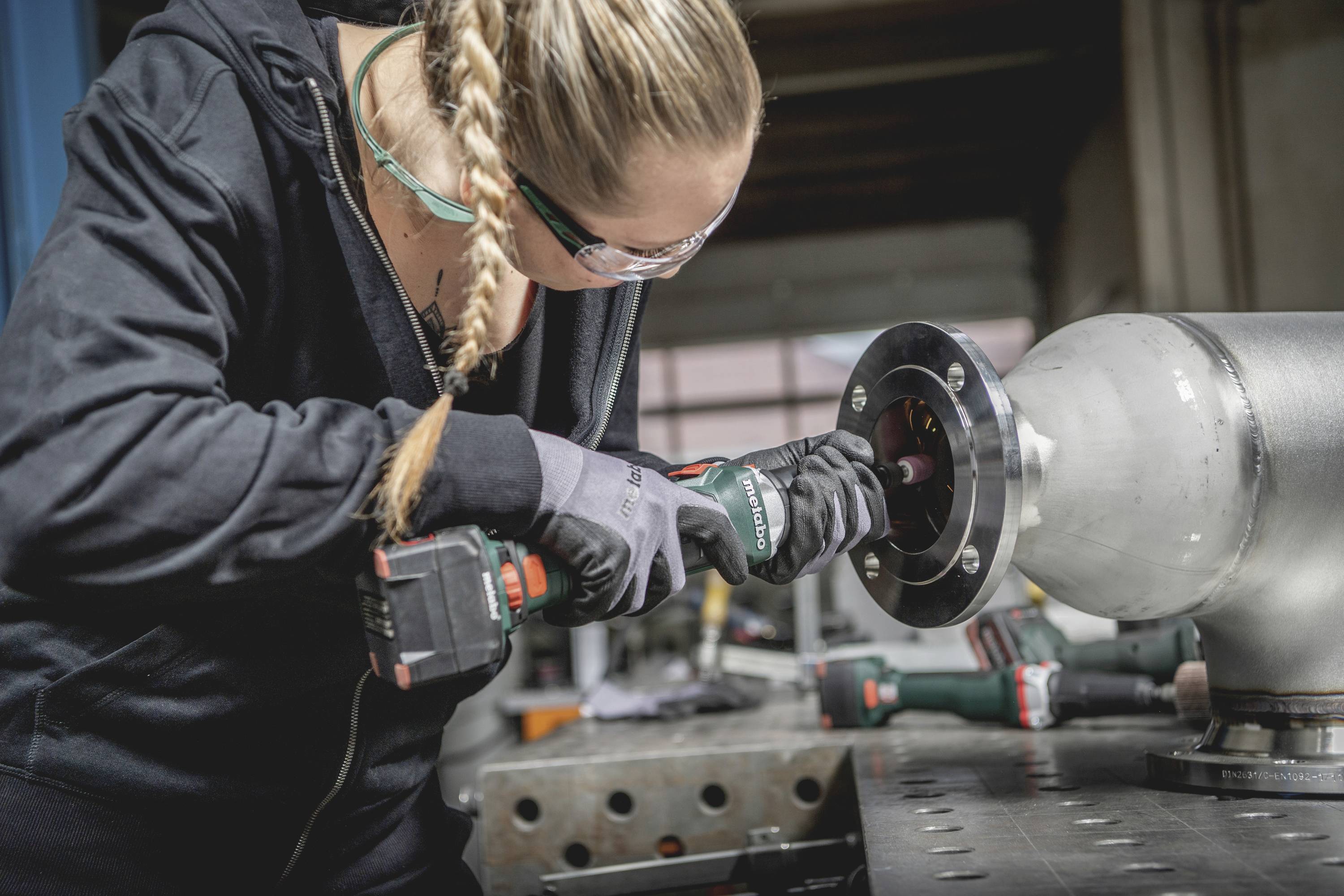 Frau in Schutzbrille und Handschuhen arbeitet konzentriert mit einer Bohrmaschine an einem Metallteil in einer Werkstattumgebung.