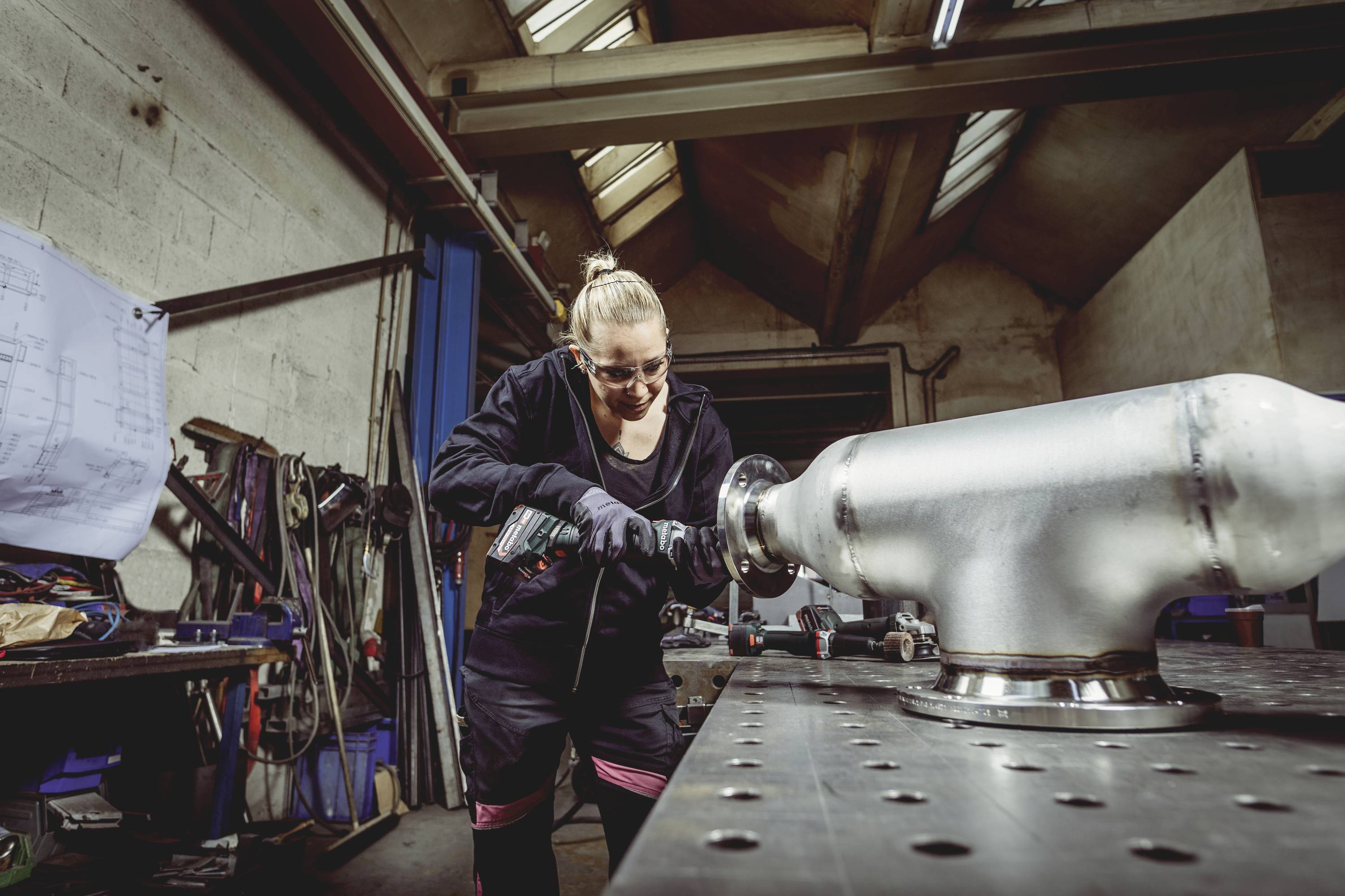 Frau in Werkstatt bearbeitet Metallwerkstück mit Elektrowerkzeug. Im Hintergrund sind Werkzeuge und Baupläne sichtbar.