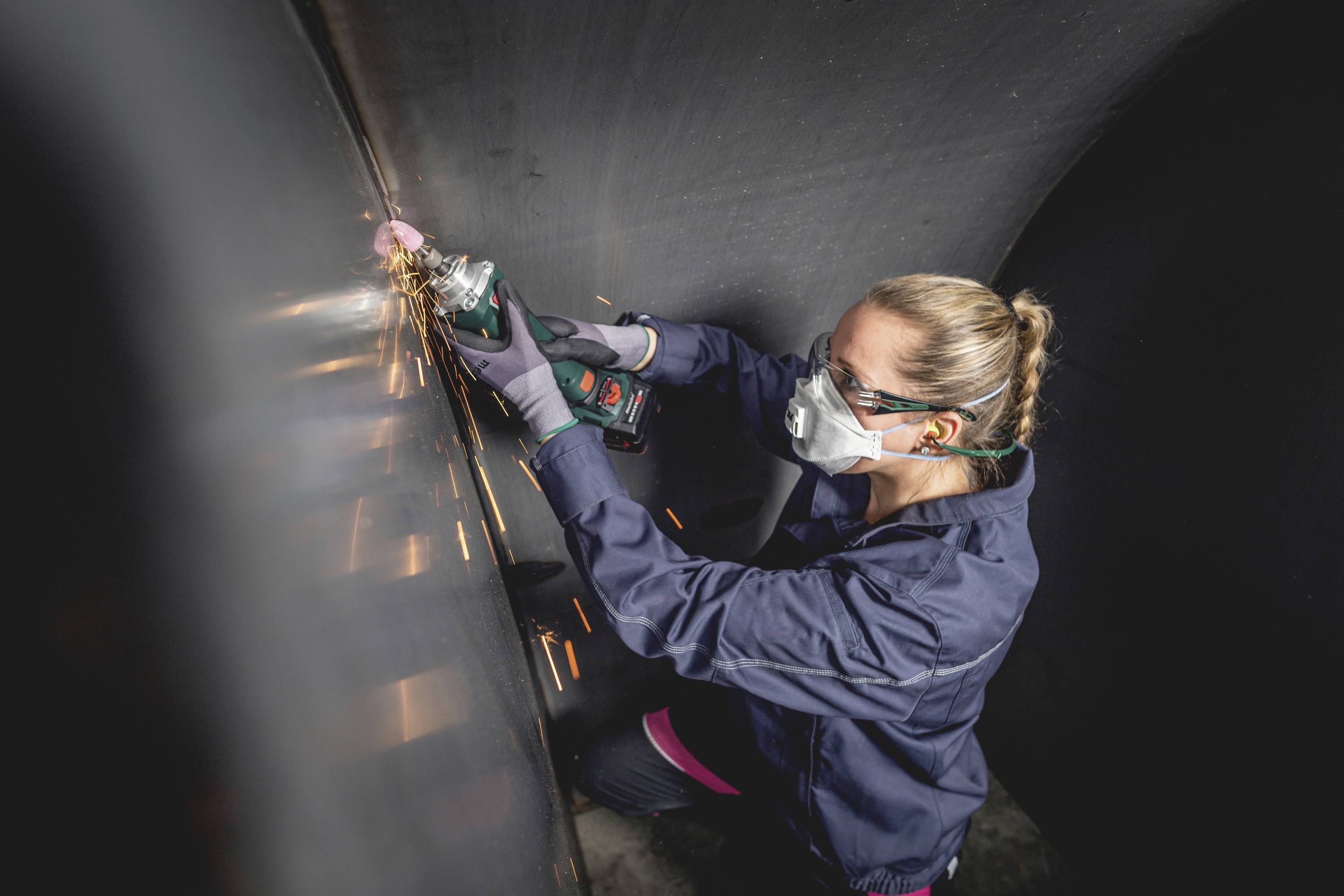 Eine Person mit Schutzbrille und Maske schleift eine Metalloberfläche mit einem Elektrowerkzeug, Funken sprühen dabei.