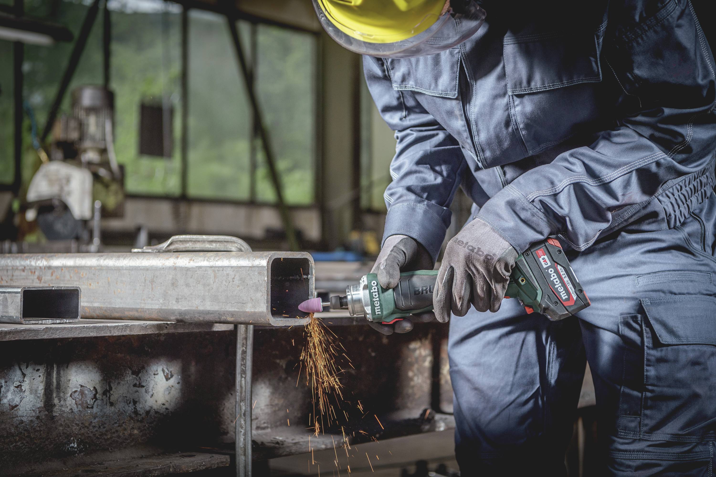 Eine Person in Arbeitskleidung schleift Metall mit einem Winkelschleifer. Funken fliegen. Die Szene findet in einer Werkstatt statt.