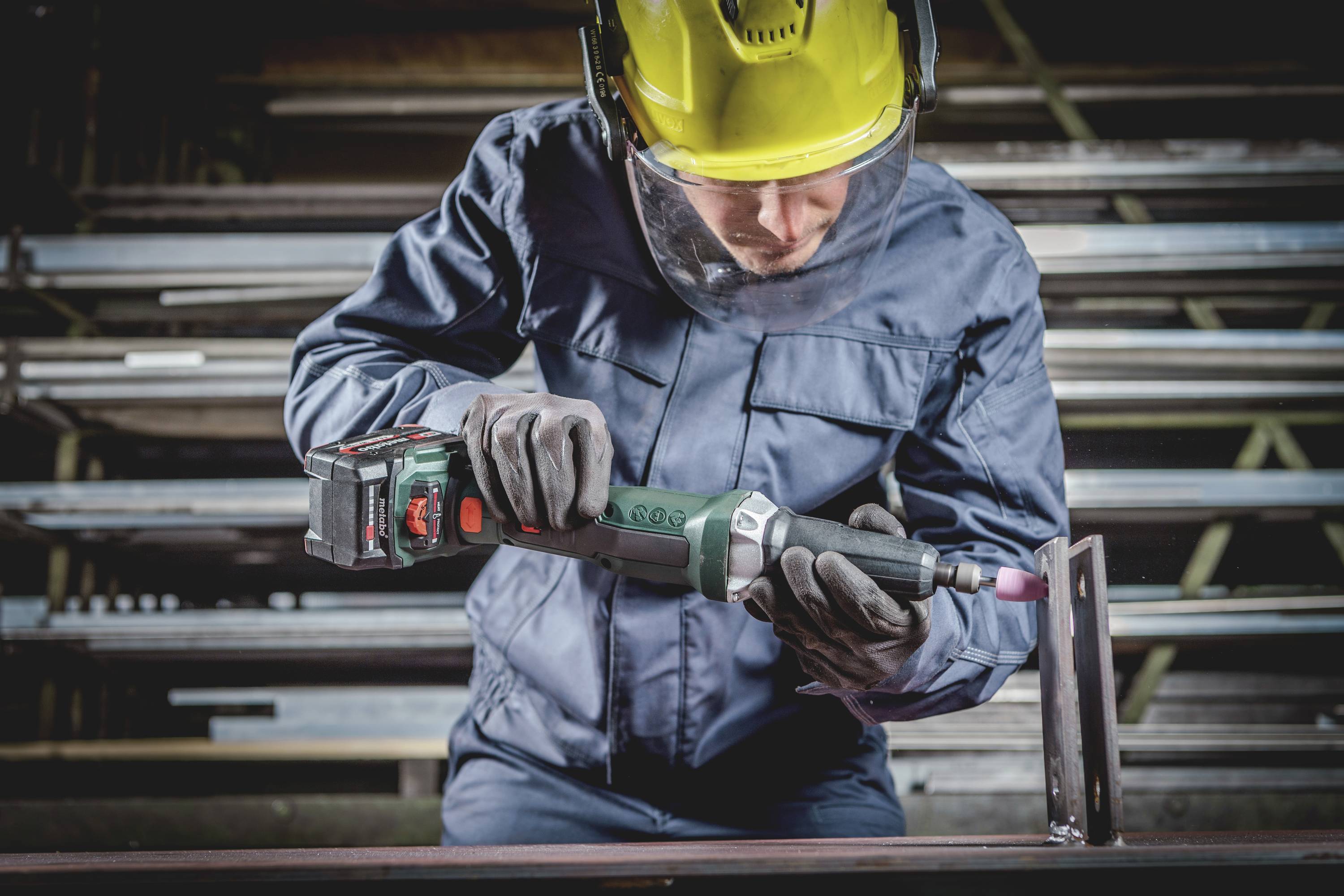 Ein Arbeiter in Schutzkleidung und Helm verwendet ein Elektrowerkzeug zum Schleifen von Metall in einer Werkstatt.
