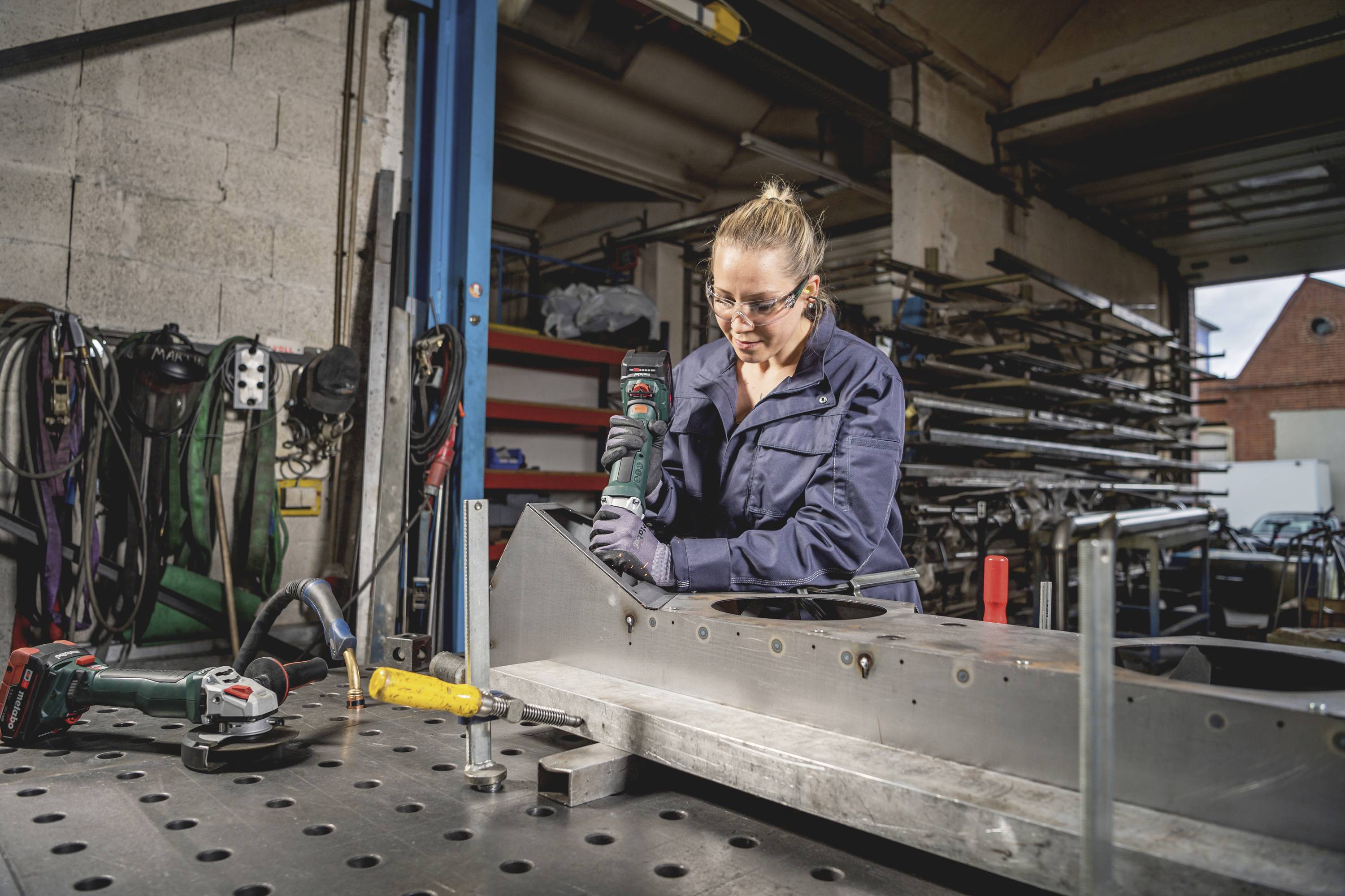 Eine Frau arbeitet in einer Werkstatt und bohrt mit einem Akkuschrauber in ein Metallstück. Im Hintergrund sind Werkzeuge zu sehen.