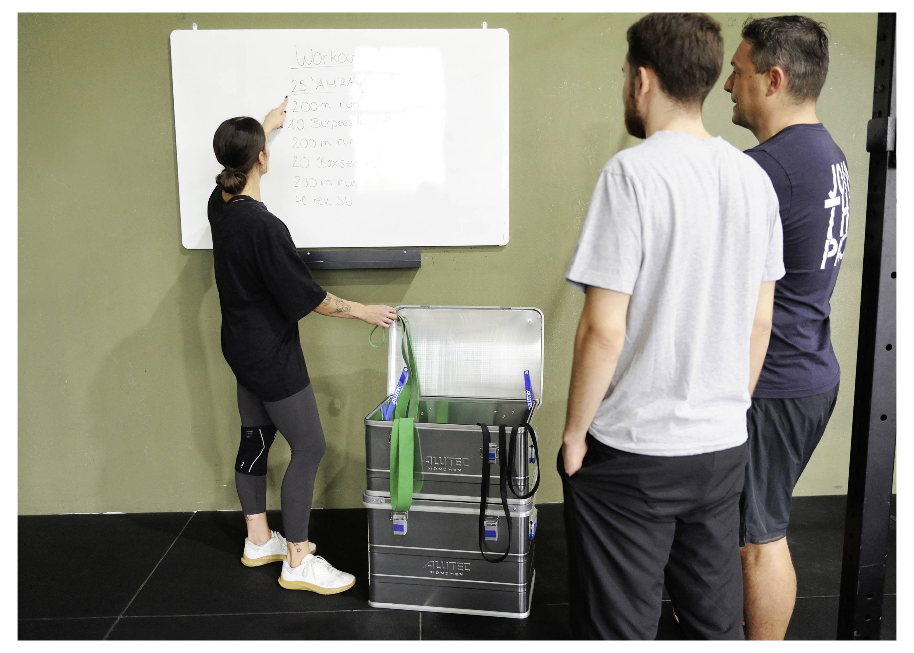 Eine Frau zeigt auf eine Liste auf einem Whiteboard, während drei Männer in der Nähe stehen, neben offenen Ausrüstungskisten, in einer Fitnessstudio-Umgebung.