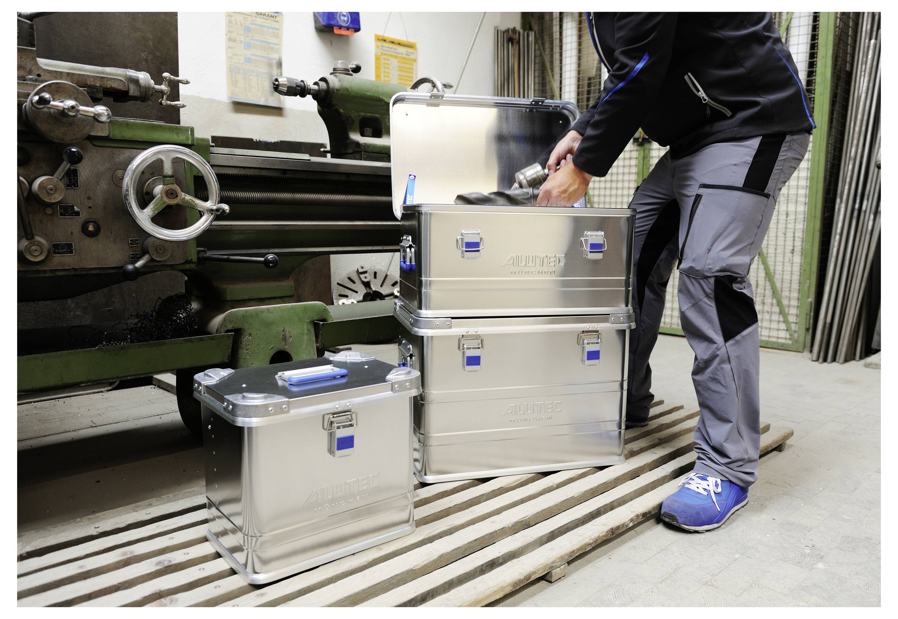 Person, die Metalllagerkästen in einer Werkstatt neben einer großen Industriemaschine stapelt, organisiert Werkzeuge und Ausrüstung.