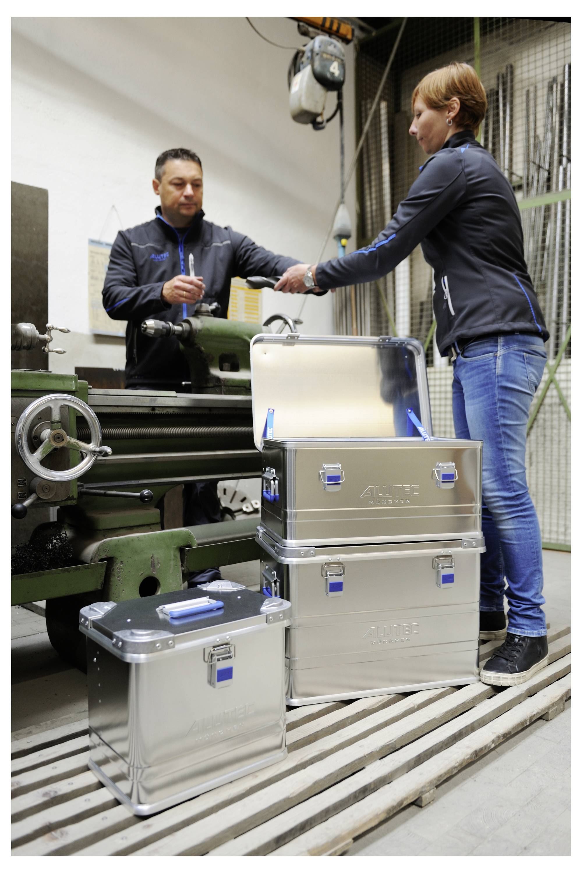 Ein Mann und eine Frau stehen in einer Werkstatt neben einer Metallmaschine und schauen sich Dokumente an. Drei silberne Lagerkisten sind in der Nähe gestapelt.