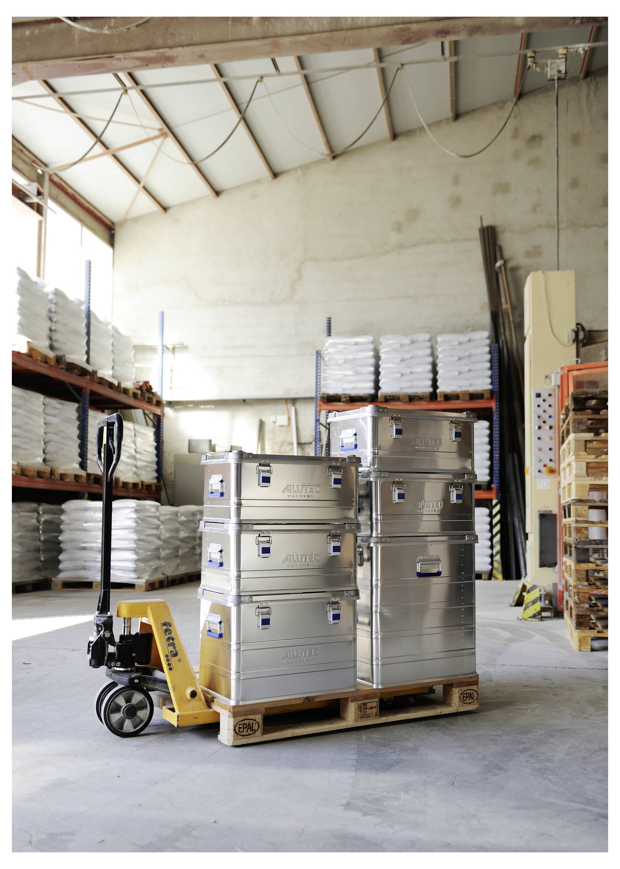 Eine Palette mit gestapelten Metalllagerbehältern in einem Lagerhaus. Regale mit verpackten Waren säumen die Wände, mit einer Handkarre auf der linken Seite.