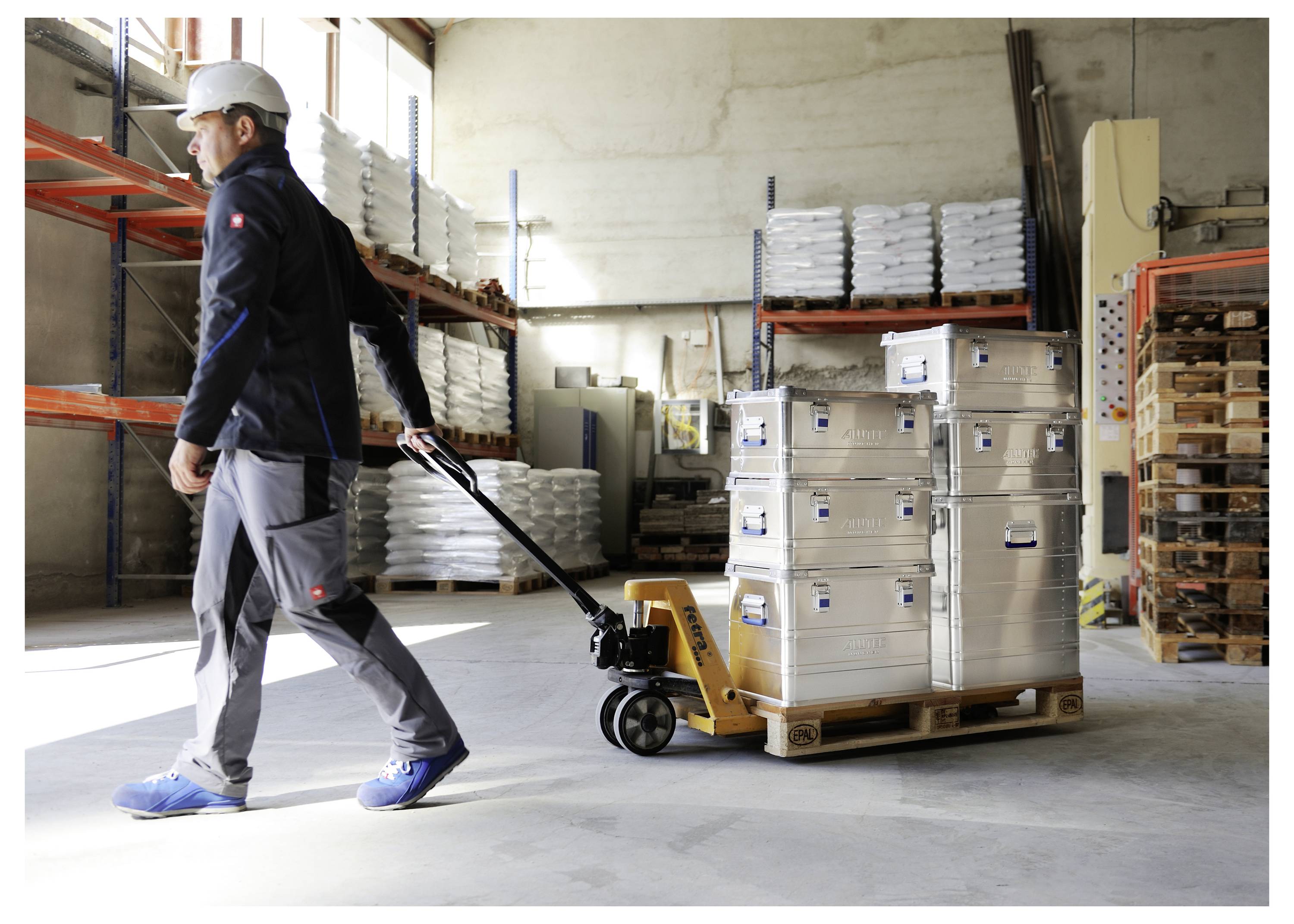 Ein Arbeiter in einem Lagerhaus zieht einen beladenen Hubwagen mit gestapelten Metallkisten. Regale mit zusätzlichen Kisten befinden sich im Hintergrund.