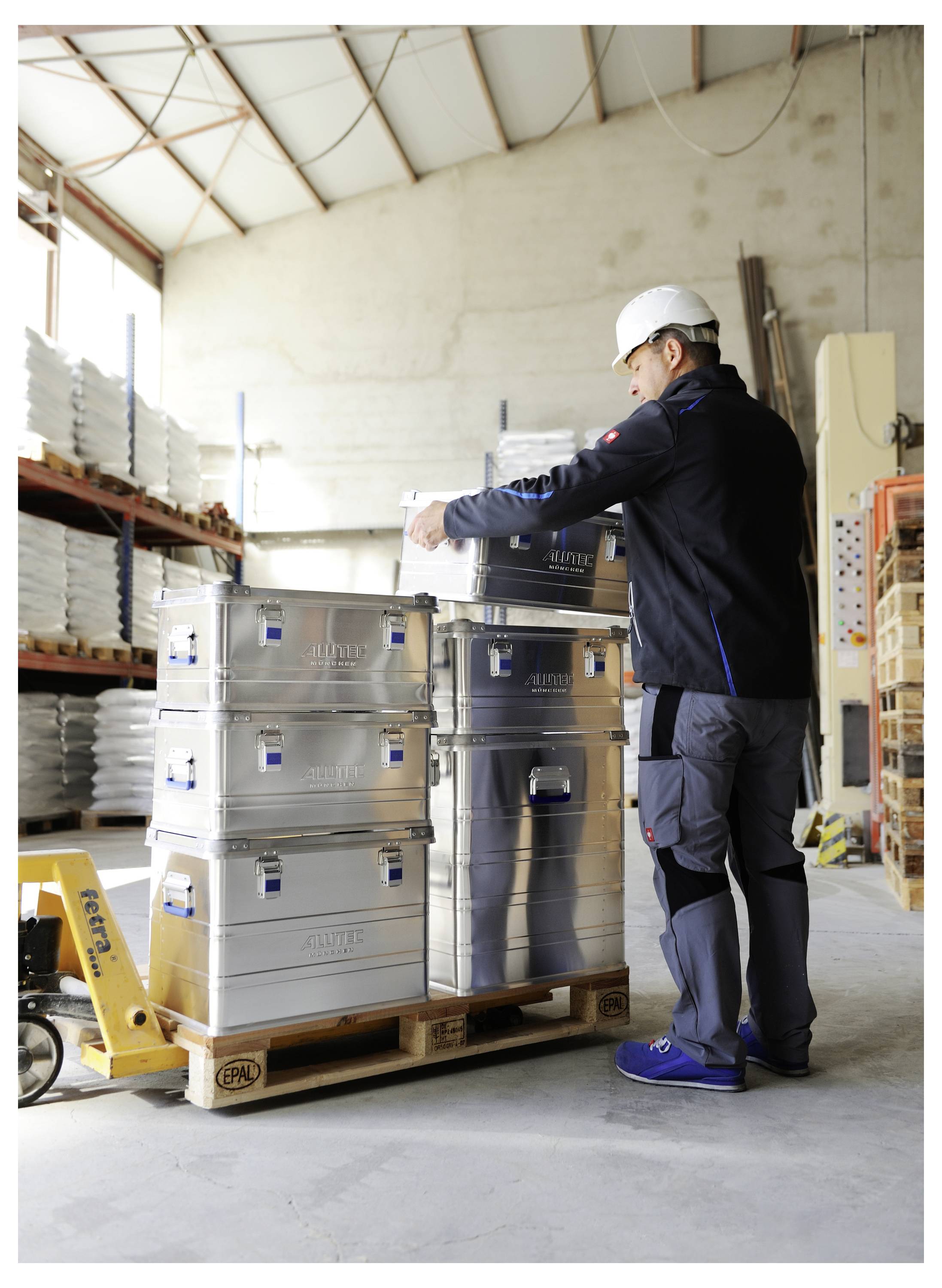 Eine Person in Schutzhelm und Arbeitskleidung untersucht gestapelte Metallkisten auf einer Palette in einer Lagerumgebung.