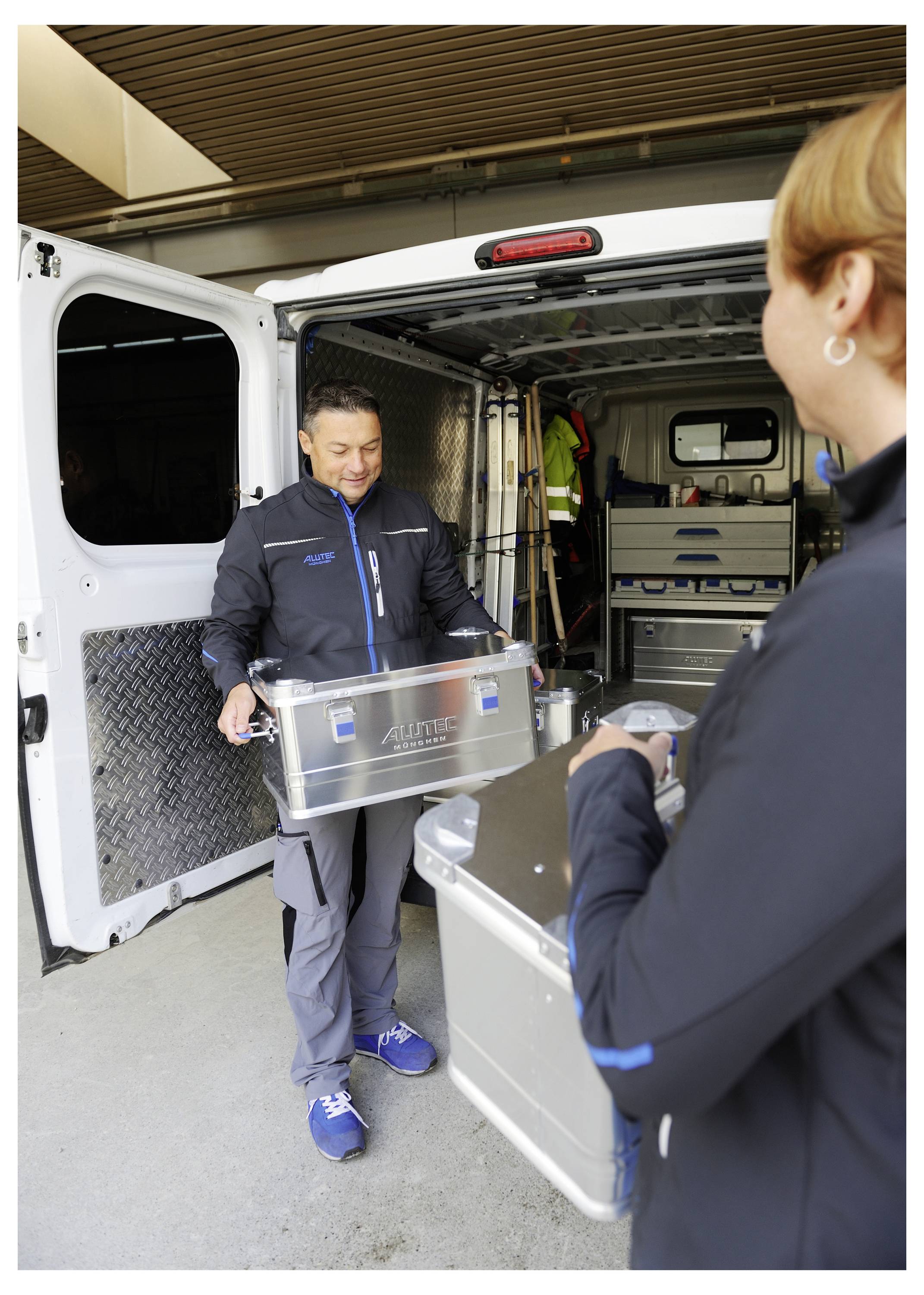 Zwei Personen laden metallische Kisten in die Rückseite eines Lieferwagens, was auf eine Transport- oder Lieferaufgabe hindeutet. Die Türen des Lieferwagens sind offen.