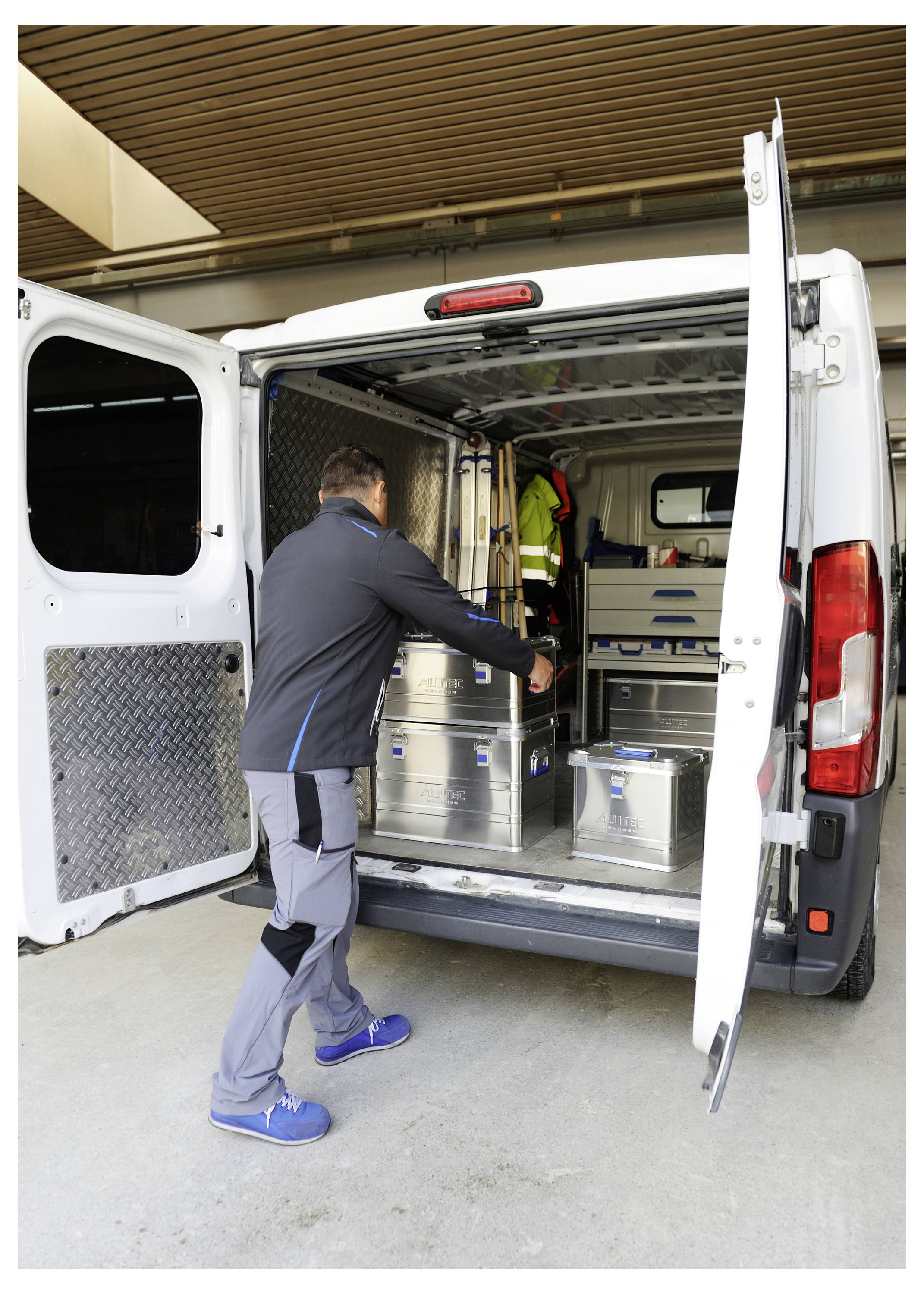 Eine Person in Arbeitskleidung lädt Metallbehälter in einen weißen Lieferwagen, der in einer Halle geparkt ist. Die Hecktüren des Wagens sind offen und zeigen einen organisierten Innenraum.