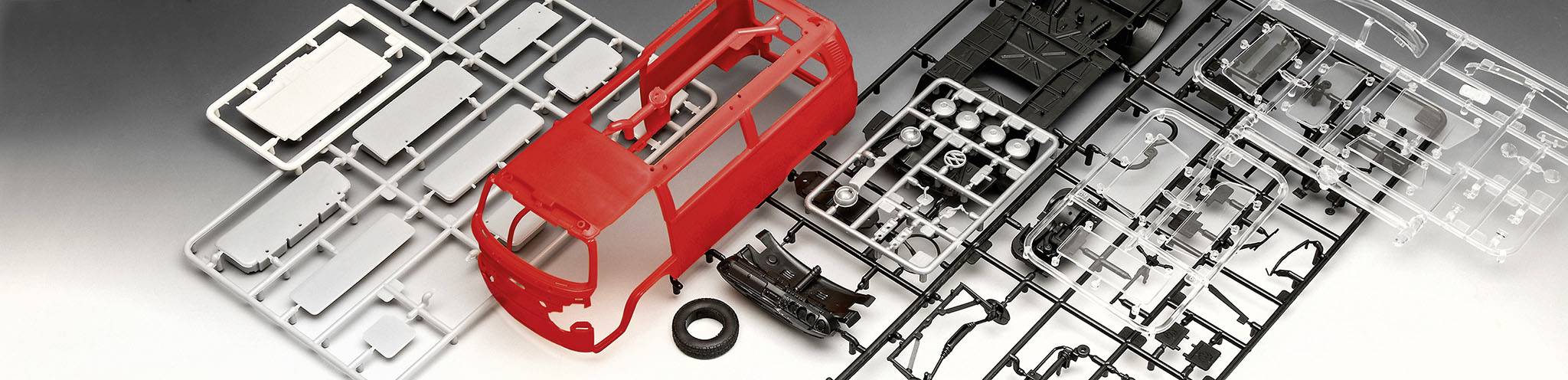 Ein Modellbausatz eines roten Kleinbusses mit verschiedenen Bauteilen und Rahmen auf einem Tisch ausgebreitet.