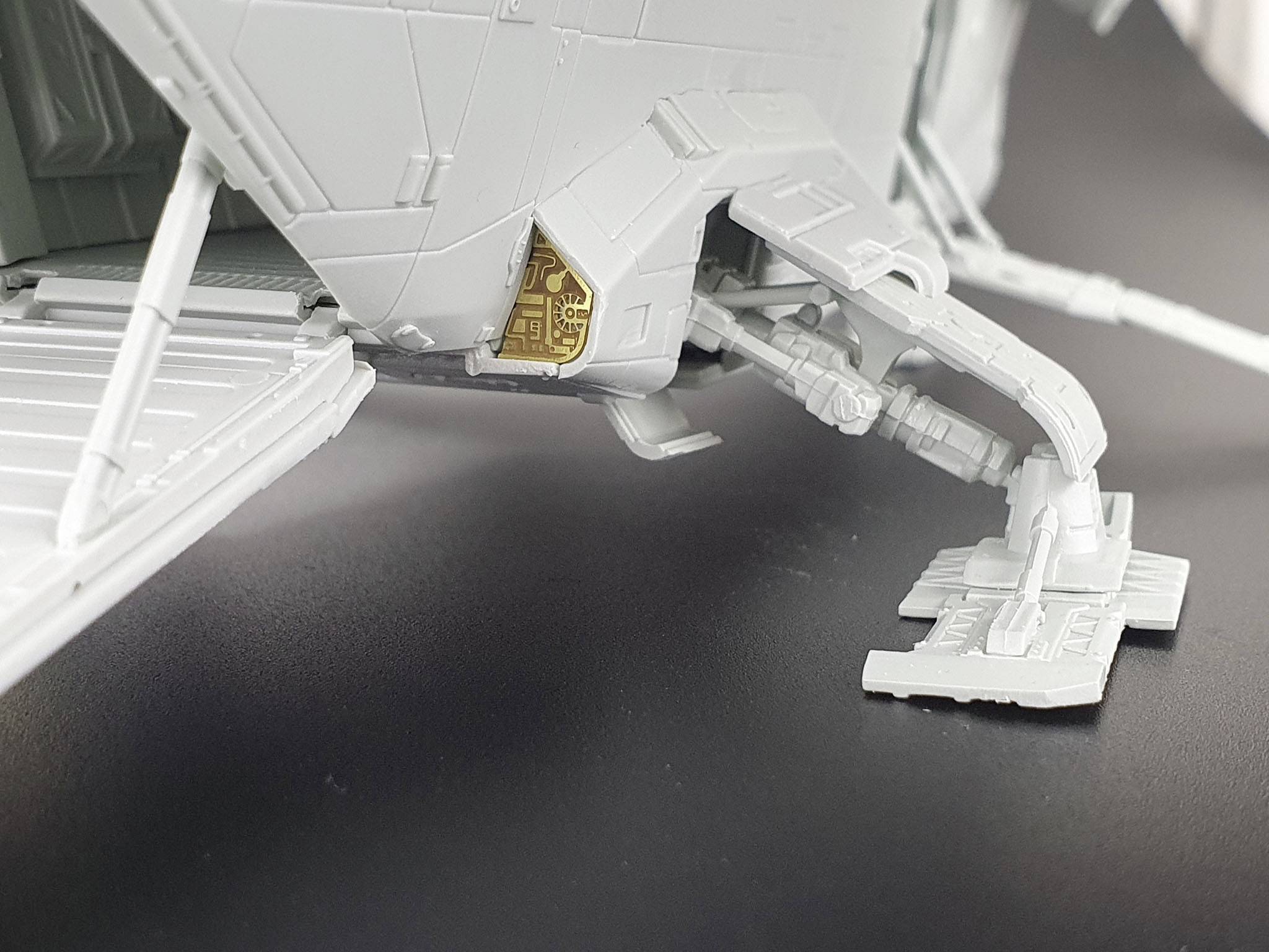 Ein graues Modell eines futuristischen Raumfahrzeugs auf einer schwarzen Oberfläche, zeigt ein komplexes Fahrwerksdetail mit goldener Komponente.