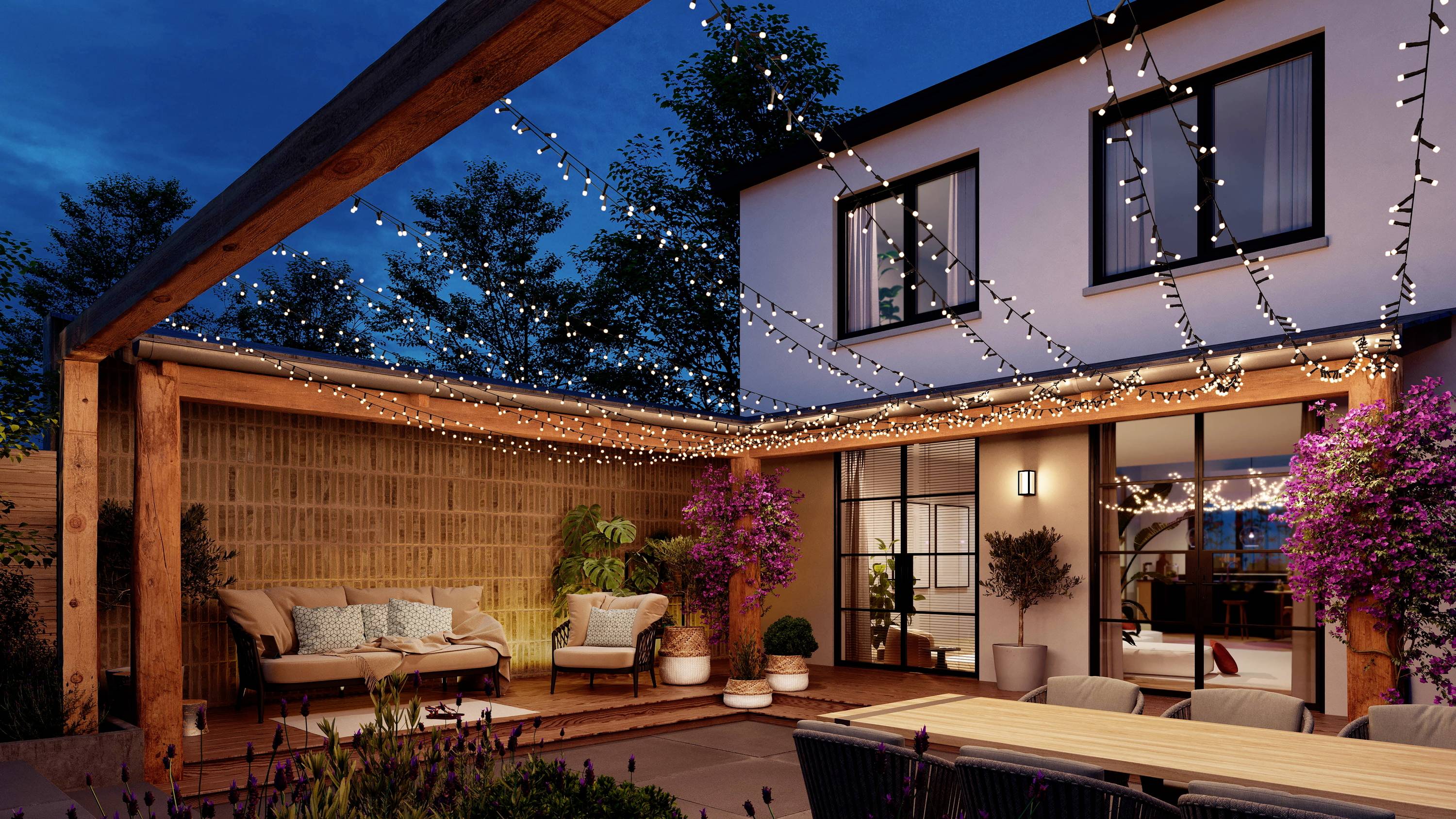 Moderne Terrasse bei Nacht, geschmückt mit Lichterketten. Sitzbereich mit Sofa und Pflanzen unter Holzpergola neben beleuchtetem Haus.