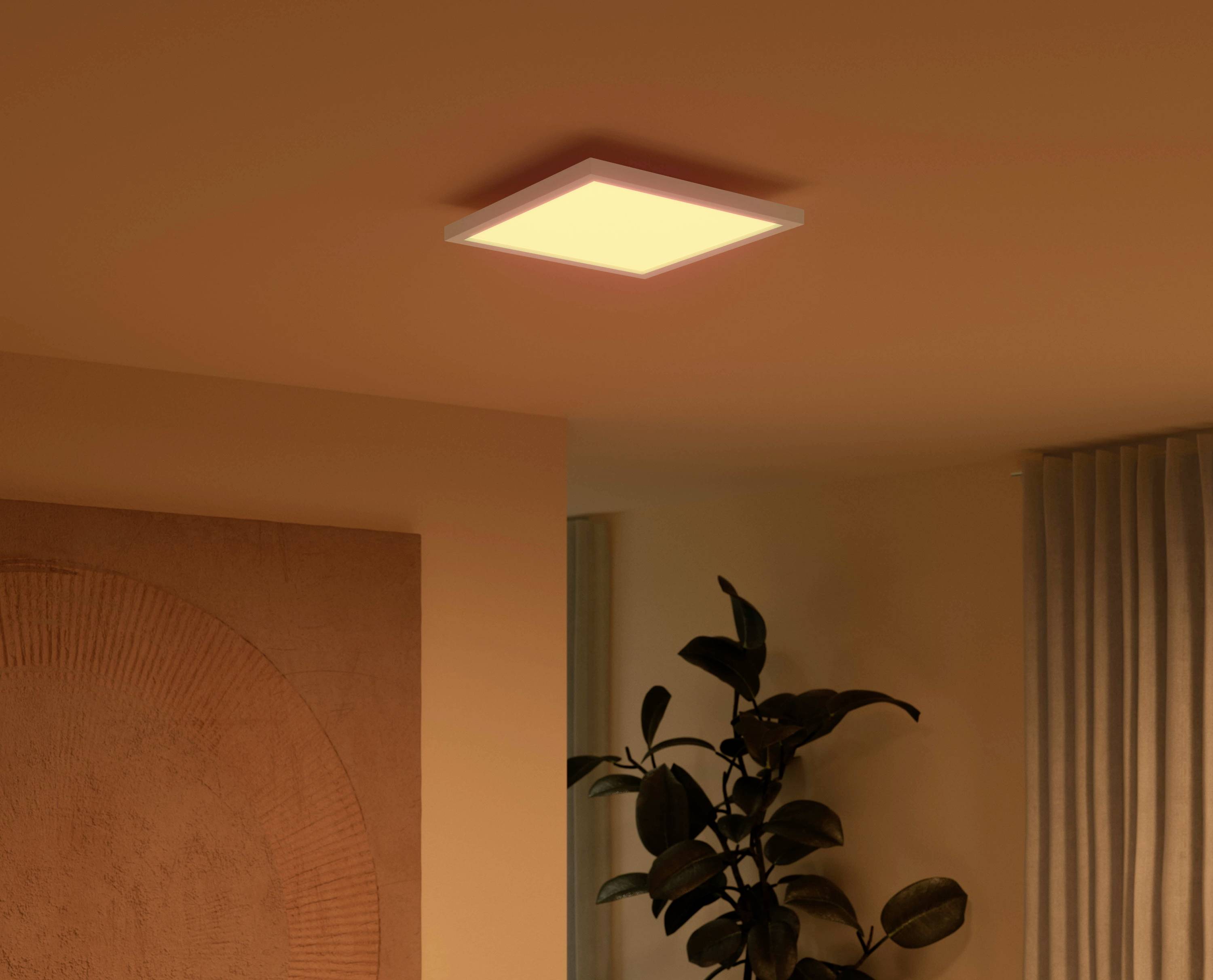 Eine quadratische Deckenleuchte leuchtet warmweiß in einem gemütlichen Raum mit einer Pflanze und Vorhängen im Hintergrund.