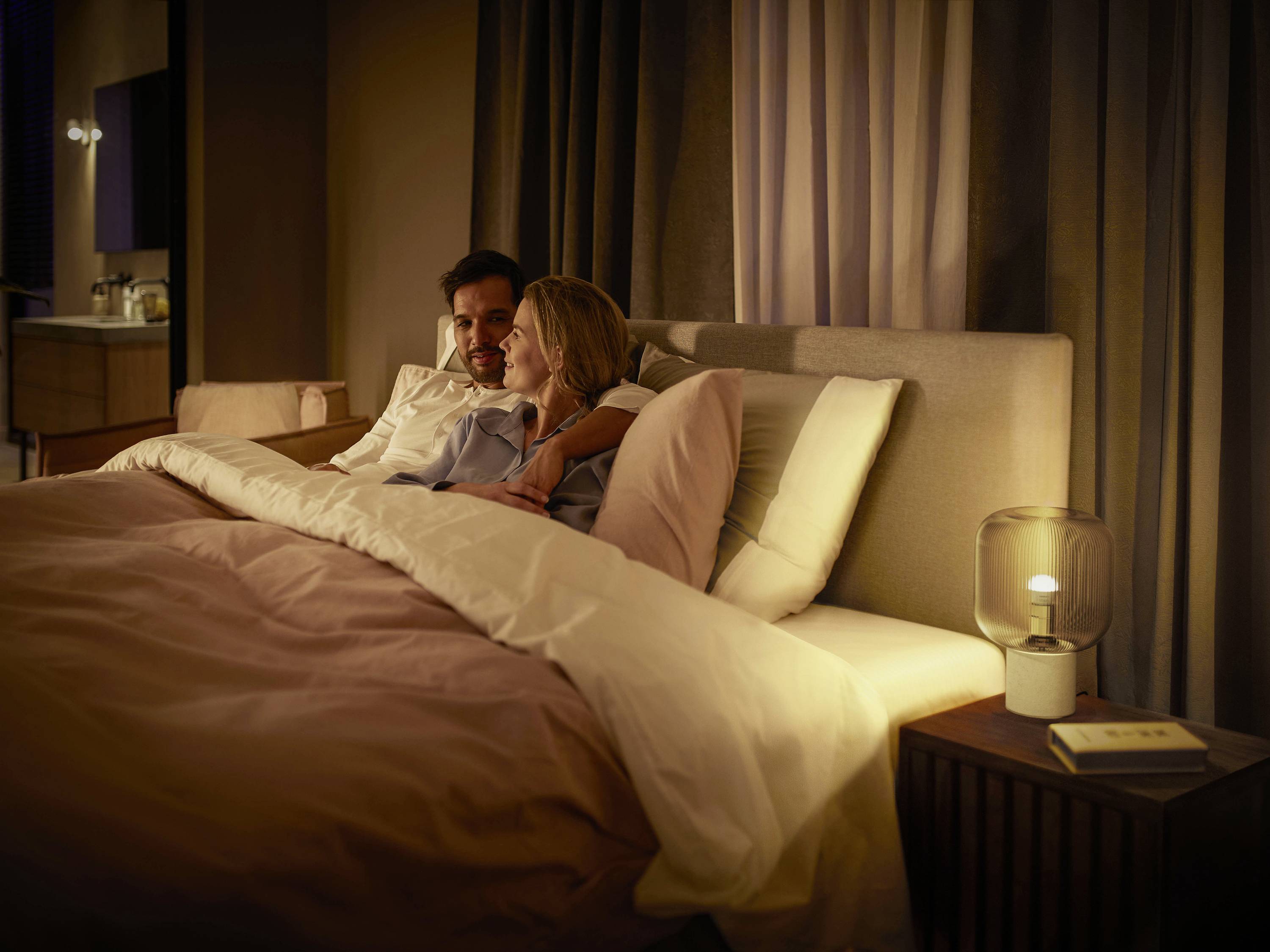 Ein Paar liegt in einem gemütlichen Bett in einem sanft beleuchteten Schlafzimmer. Ein Buch und eine Lampe stehen auf dem Nachttisch.