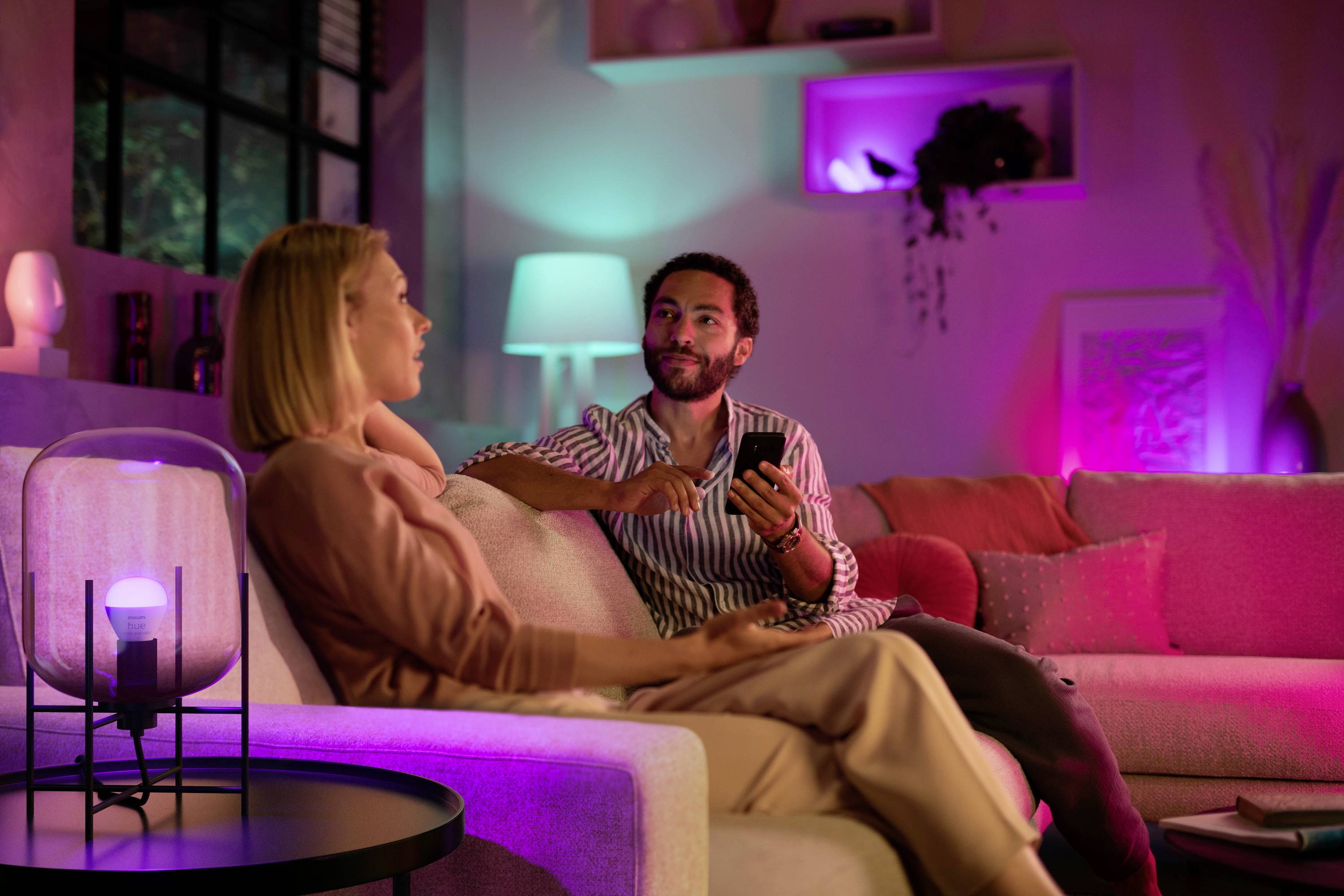 Eine Frau und ein Mann sitzen auf einem Sofa in einem abgedunkelten Raum mit bunten Lichtern. Sie schauen sich an und unterhalten sich.