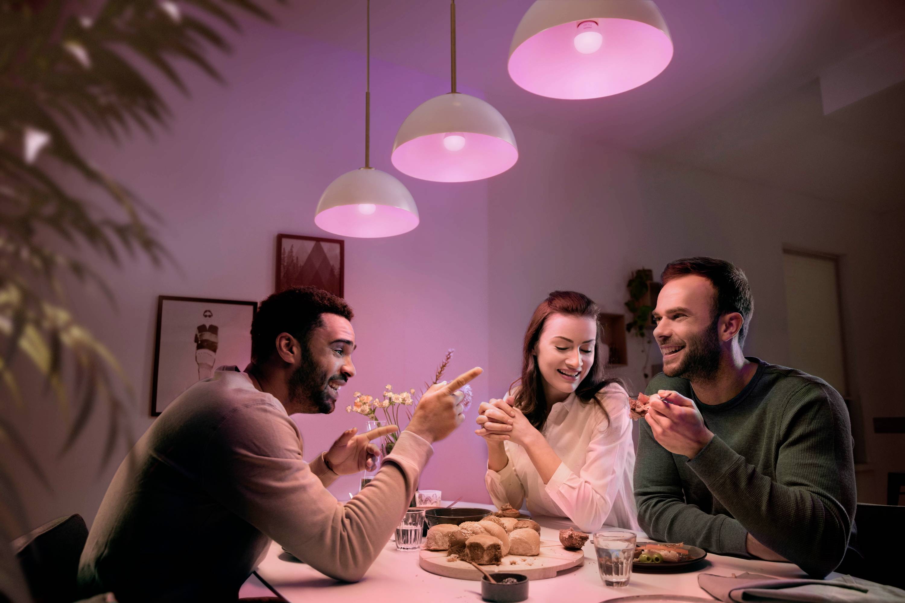 Drei Personen sitzen an einem Tisch unter rosa Leuchten, lachen und essen gemeinsam. Im Hintergrund sind Bilder und Pflanzen zu sehen.