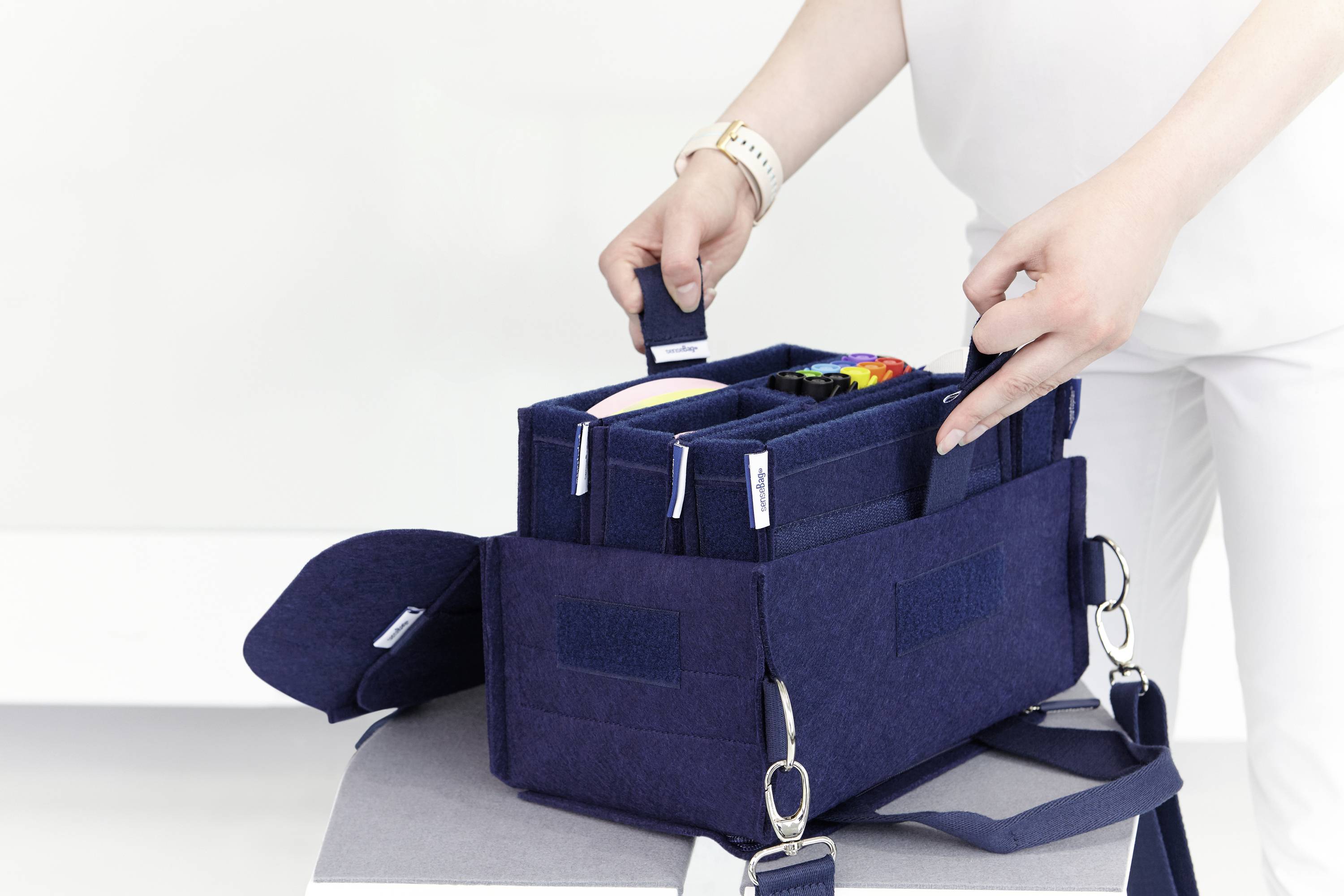 Person organisiert eine dunkelblaue Filztasche mit mehreren Fächern, die verschiedene bunte Gegenstände enthält.