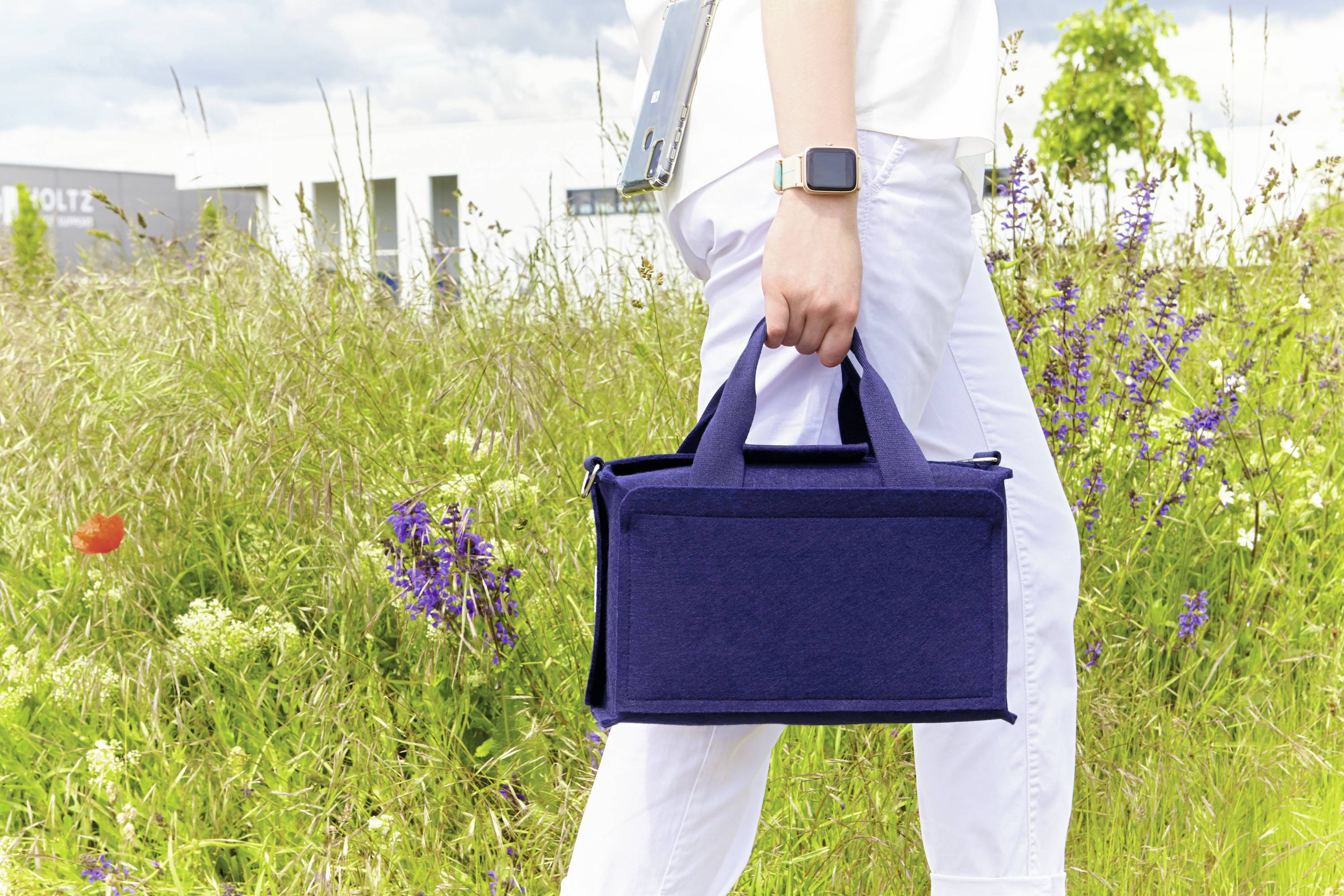 Person in weißem Outfit trägt eine blaue Tasche über einer Wiese mit Blumen im Hintergrund.