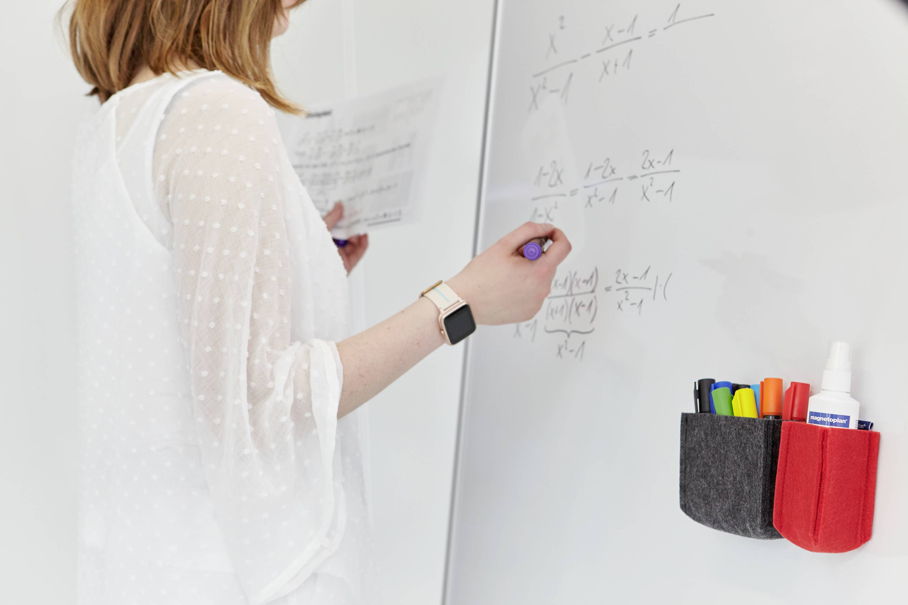 Eine Person schreibt mathematische Formeln auf ein Whiteboard. Daneben sind bunte Marker in einer Halterung zu sehen.
