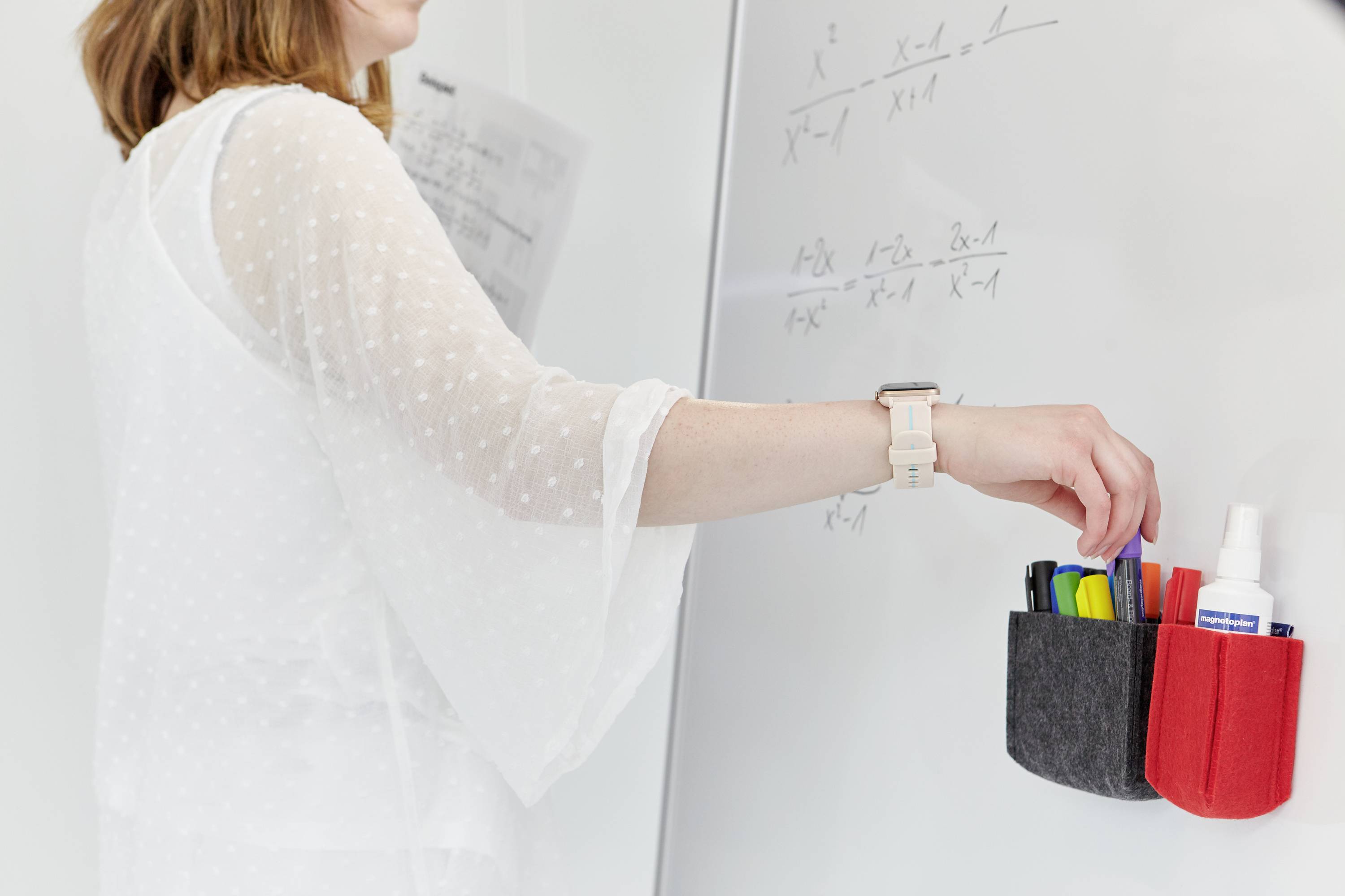Eine Person schreibt mathematische Formeln an ein Whiteboard. In der Hand hält sie Stifte und Marker aus einem Wandhalter.