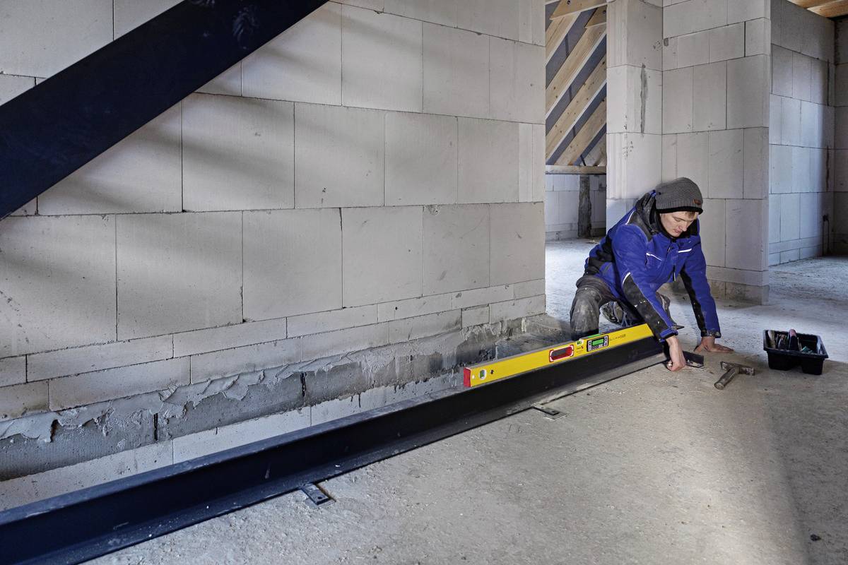 Eine Person in Arbeitskleidung nivelliert mit einer Wasserwaage einen Stahlträger in einem Rohbau. Wand und Balken im Hintergrund.