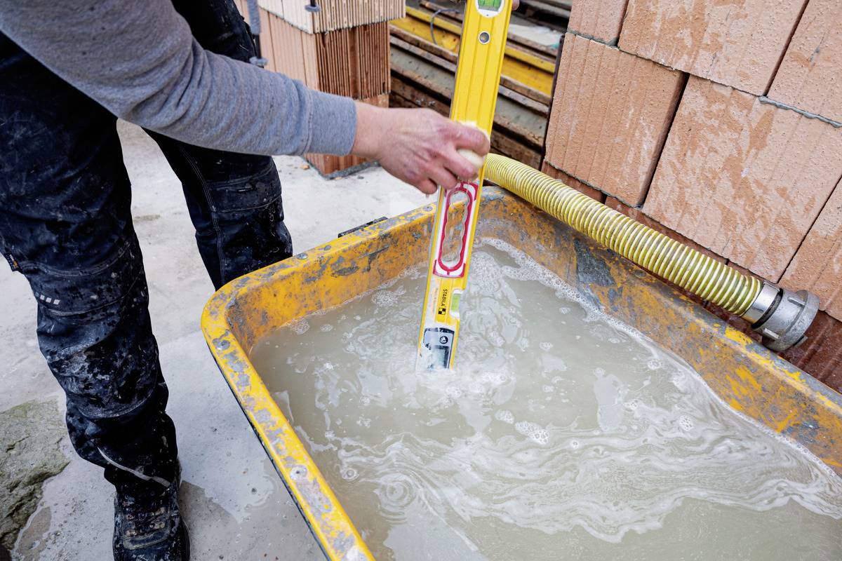 Person hält eine Wasserwaage in einem gelben Mörtelkasten mit Wasser und Zement, um den Inhalt zu mischen. Hintergrund mit Ziegeln.