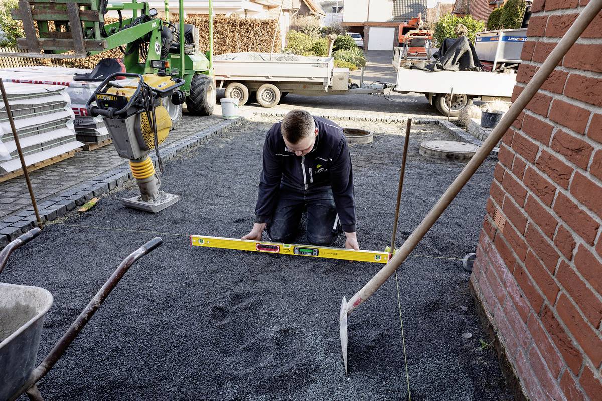 Ein Mann kniet auf einer Baustelle und nivelliert Schotter mit einer Wasserwaage. Baugeräte und Materialien sind im Hintergrund zu sehen.