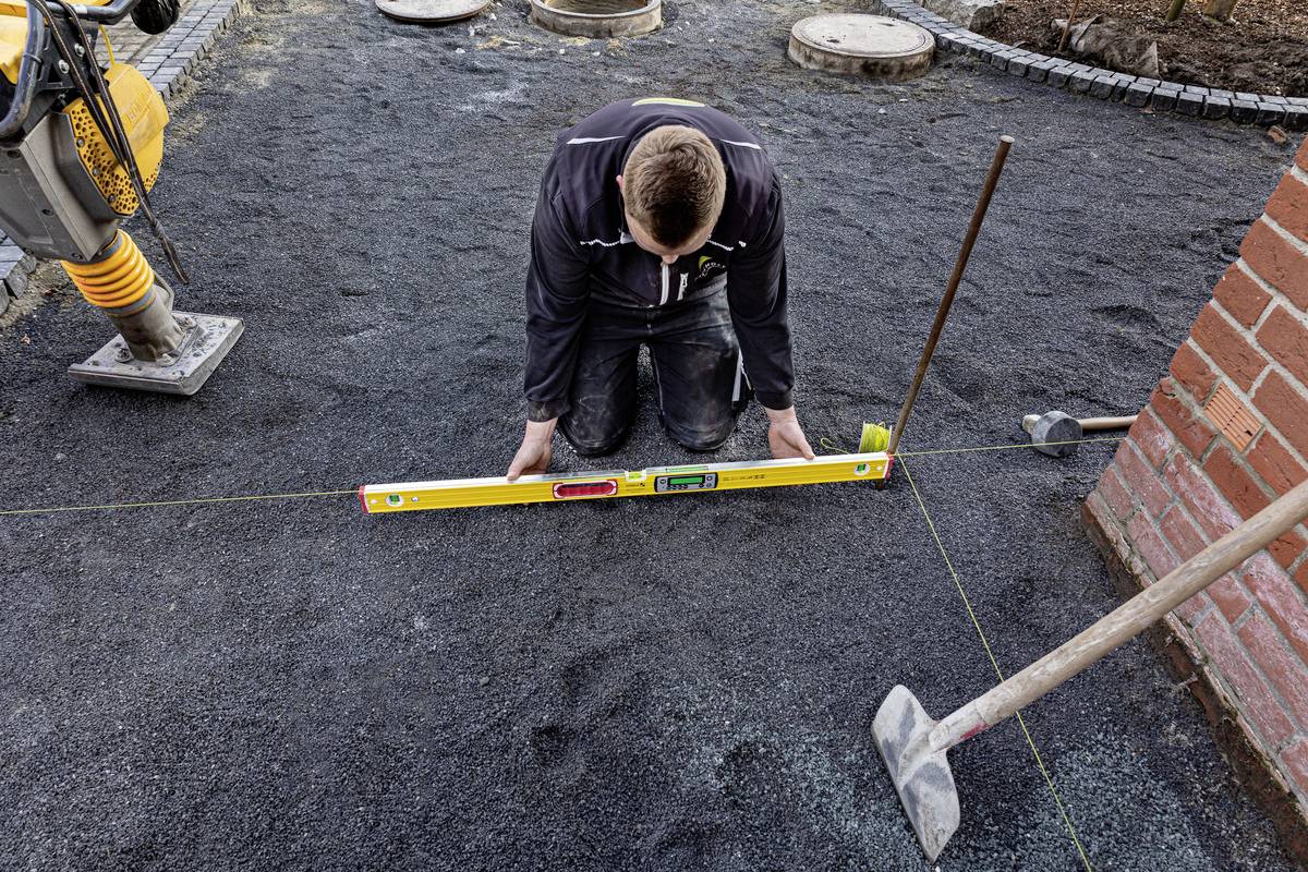 Eine Person kniet auf einer Baustelle und benutzt eine Wasserwaage, um die Ebenheit des Bodens zu prüfen. Neben der Person liegt ein Hammer.