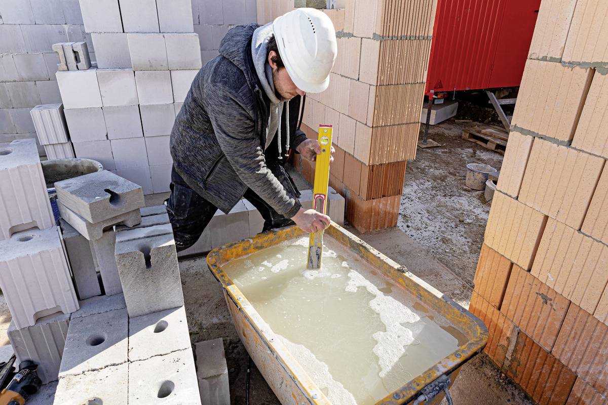 Ein Bauarbeiter prüft mit einer Wasserwaage die Ausrichtung einer gefüllten Wanne auf einer Baustelle. Stapel von Ziegeln sind im Hintergrund.