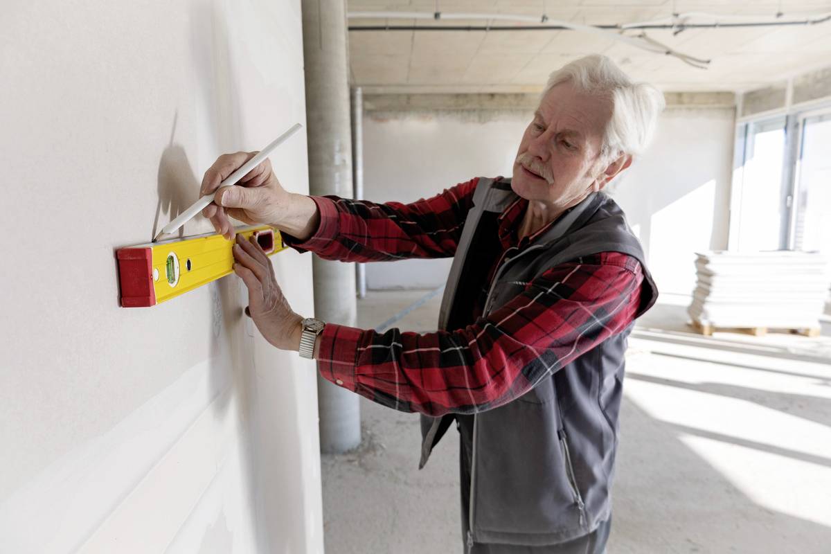 Älterer Mann mit Wasserwaage überprüft Geradlinigkeit einer Wand in einem Rohbau. Sonnenlicht fällt durch große Fenster.