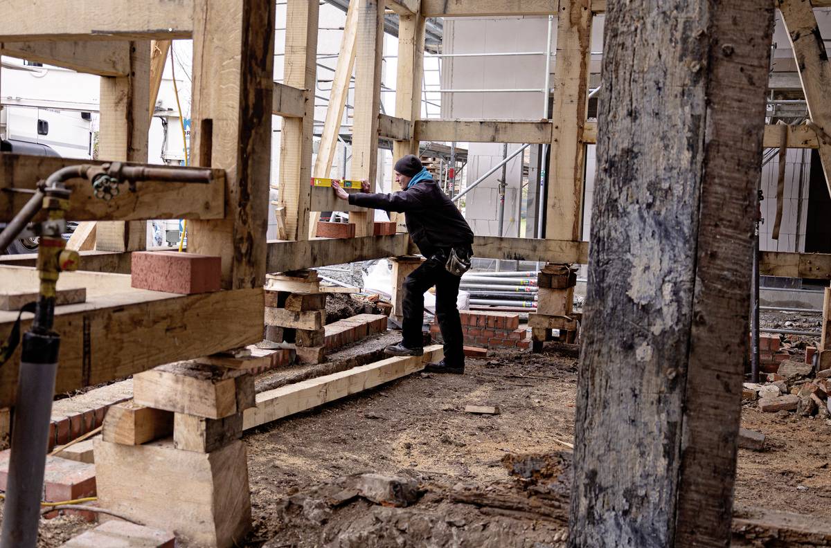 Ein Bauarbeiter in Schutzkleidung richtet ein Holzgerüst auf einer Baustelle aus. Im Hintergrund befinden sich Bauzubehör und ein Gebäude.