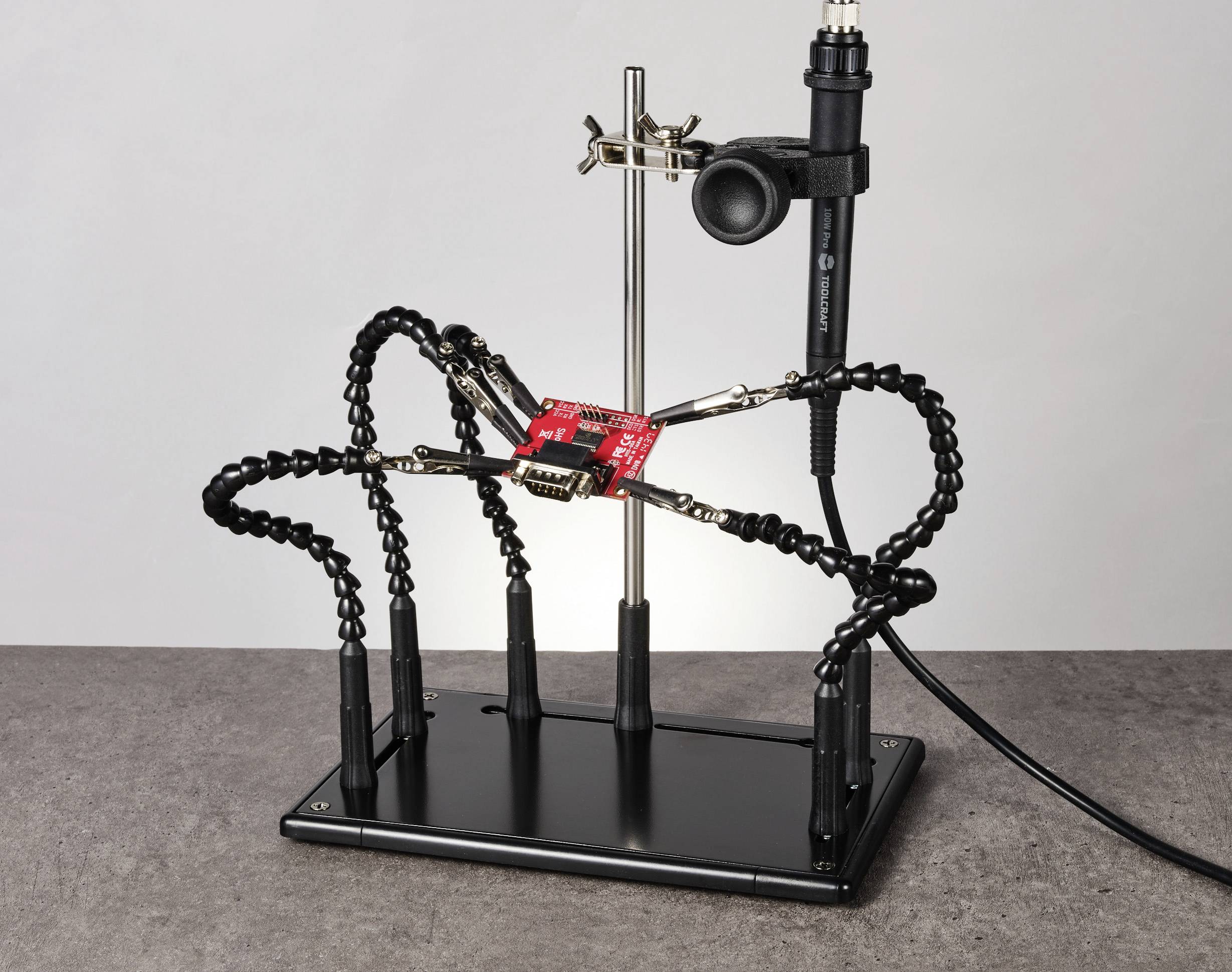 Ein Löthilfe-Werkzeug mit flexiblen Armen hält eine rote elektronische Schaltung. Ein Lötkolben ist in der Halterung befestigt.