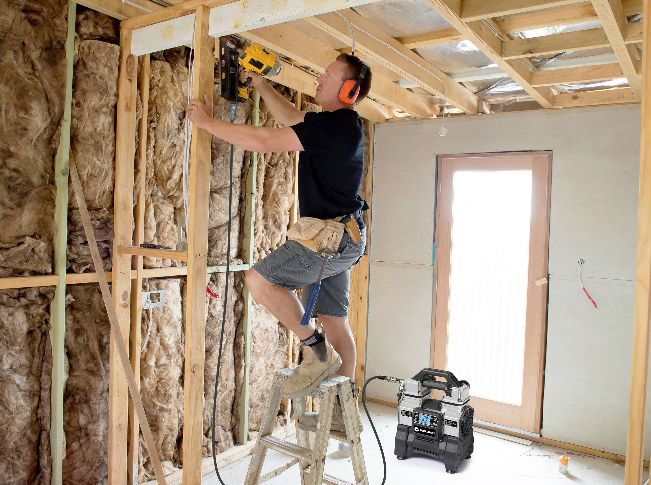 Ein Handwerker mit Schutzkopfhörern steht auf einer Leiter und bohrt in die Decke eines Holzrahmens in einem Haus im Bau.