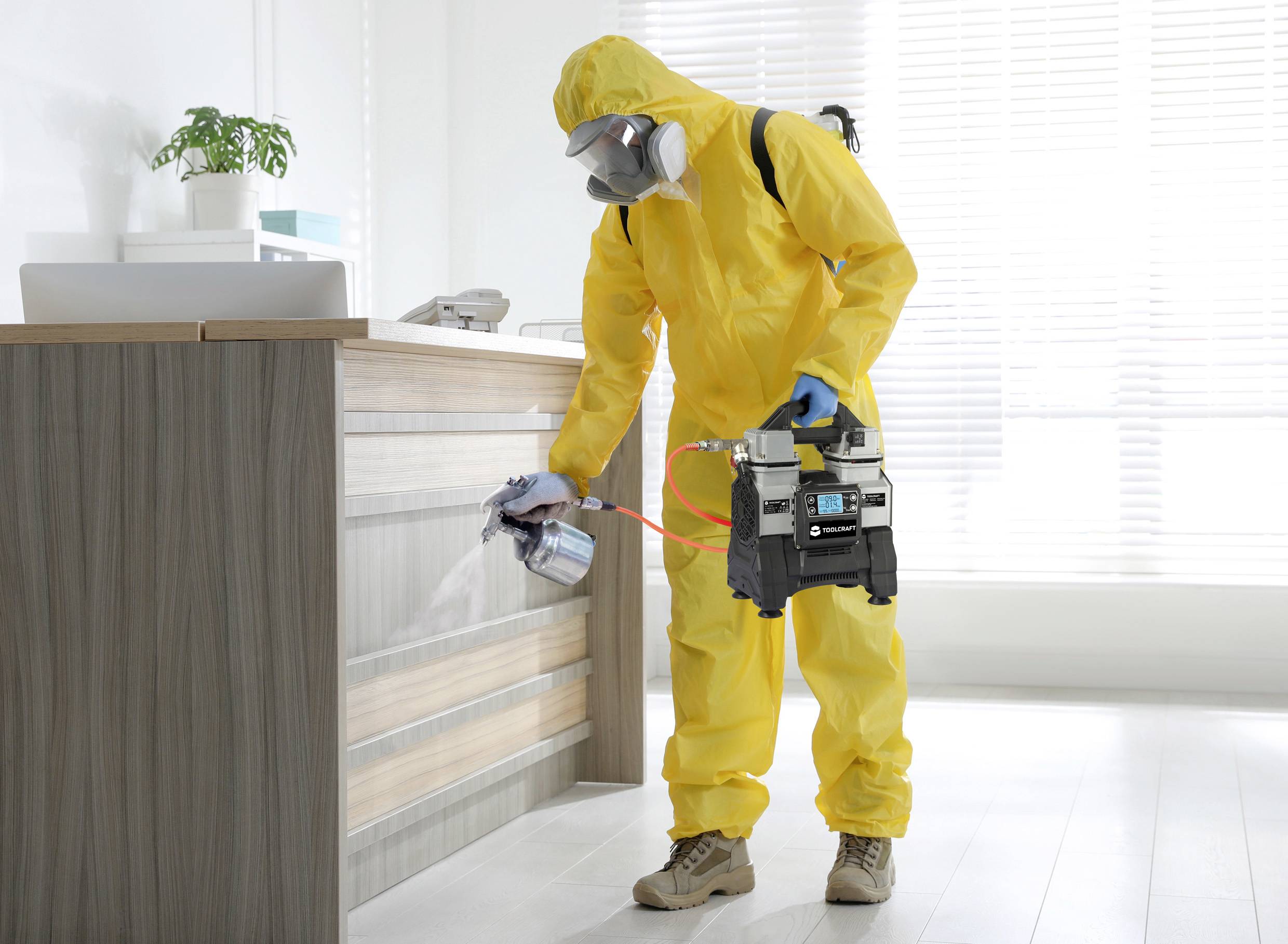 Eine Person in gelbem Schutzanzug desinfiziert eine Oberfläche mit einem Sprühgerät in einem Büro. Im Hintergrund helle Fenster.