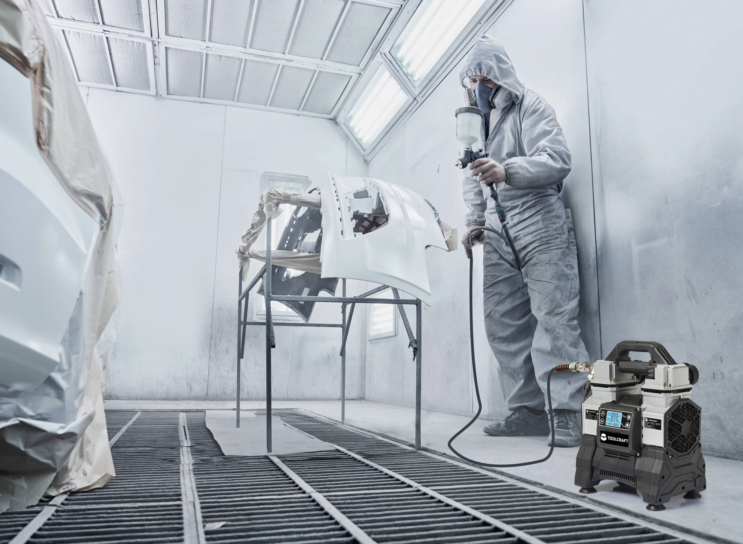 Ein Arbeiter im Schutzanzug lackiert Autoteile in einer Spritzkabine. Eine Spritzpistole und ein Luftkompressor sind sichtbar.