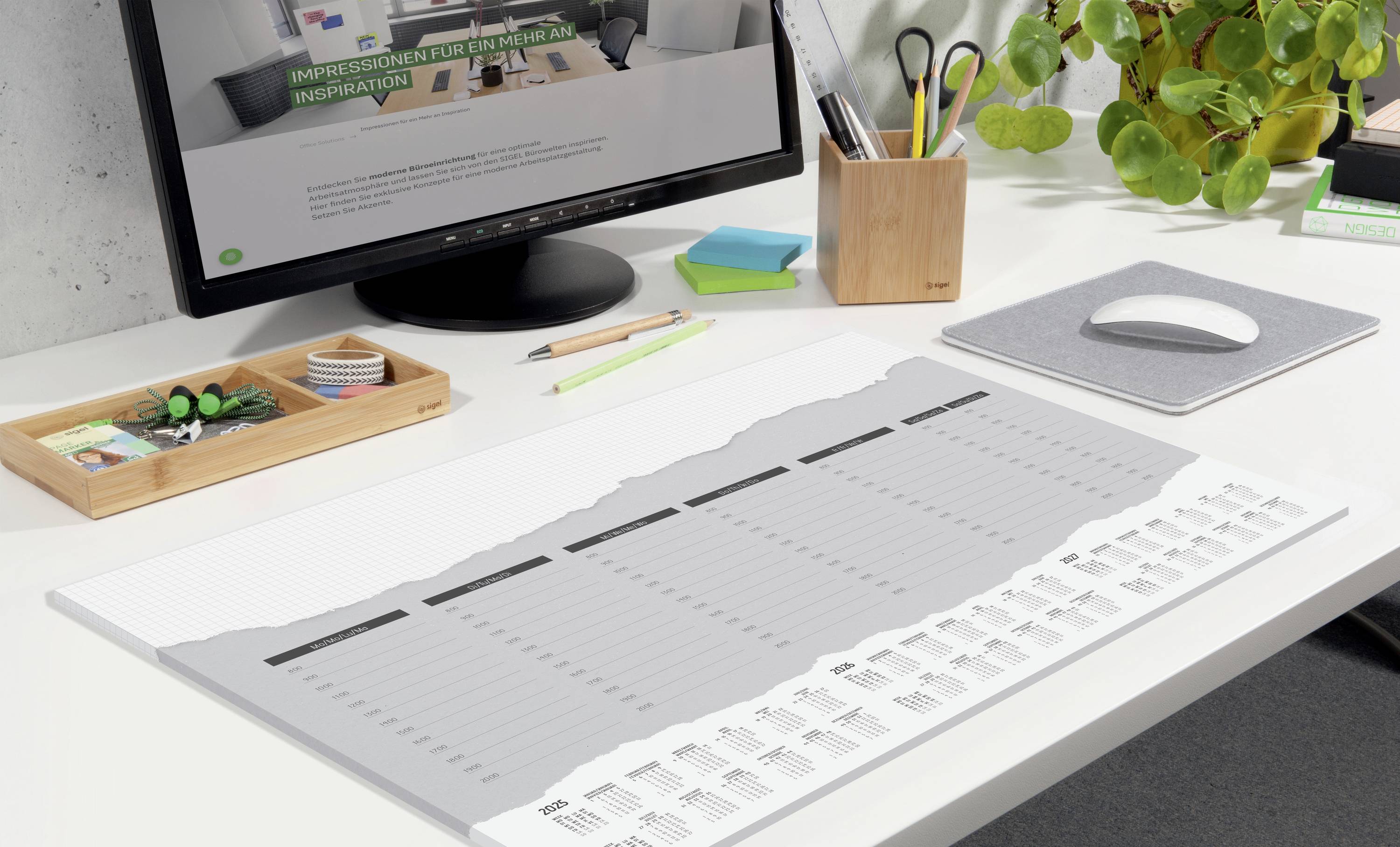 Ein aufgeräumter Schreibtisch mit einem großflächigen Kalender als Tischunterlage, umgeben von Büromaterialien und einem Monitor.
