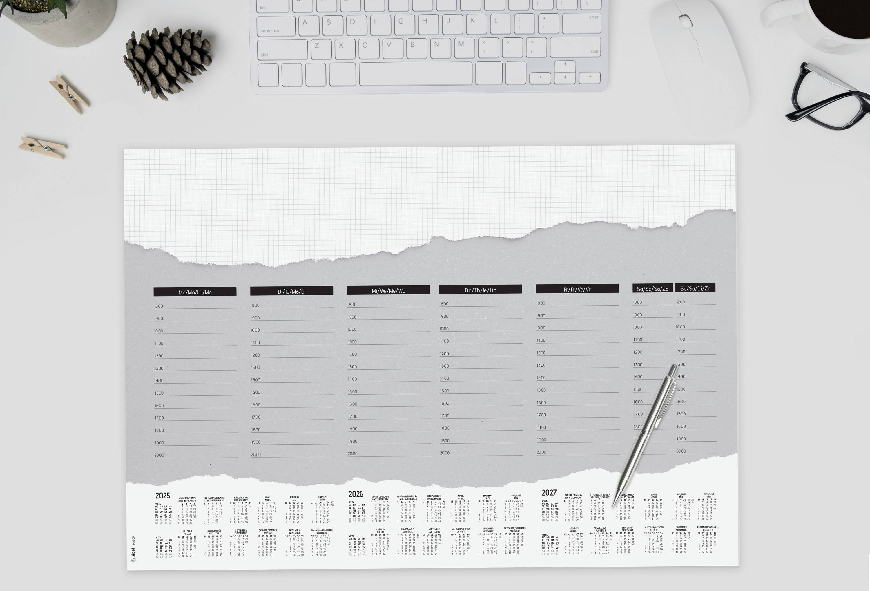 Schreibtischansicht mit Kalender-Notizblock, Stift, Tastatur, Maus und Kaffeetasse. Der Notizblock zeigt eine Monatswochenansicht.