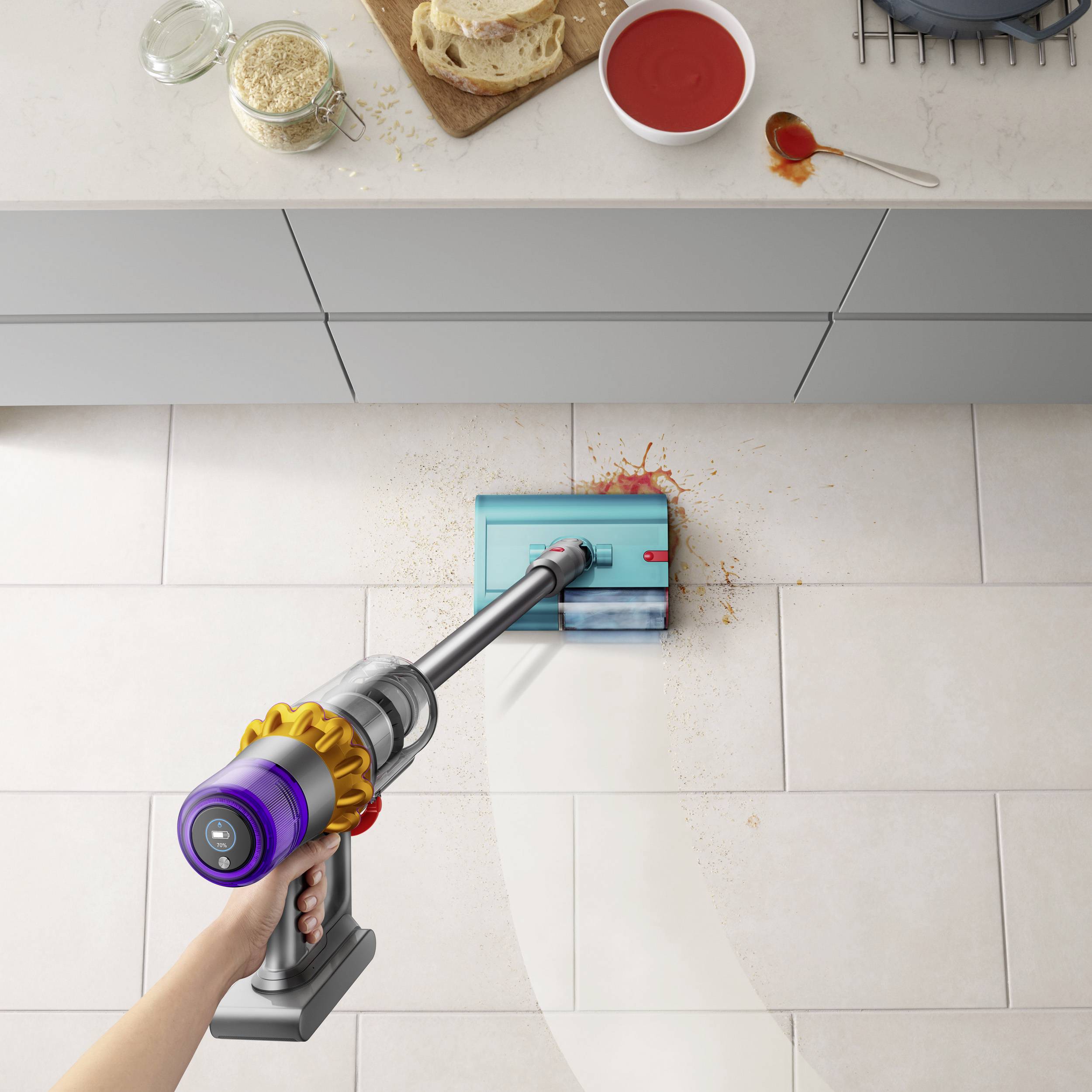 Staubsauger, der Tomatensauce auf Bodenfliesen reinigt, während Brot und Glas mit Hafer auf der Küchenzeile stehen.