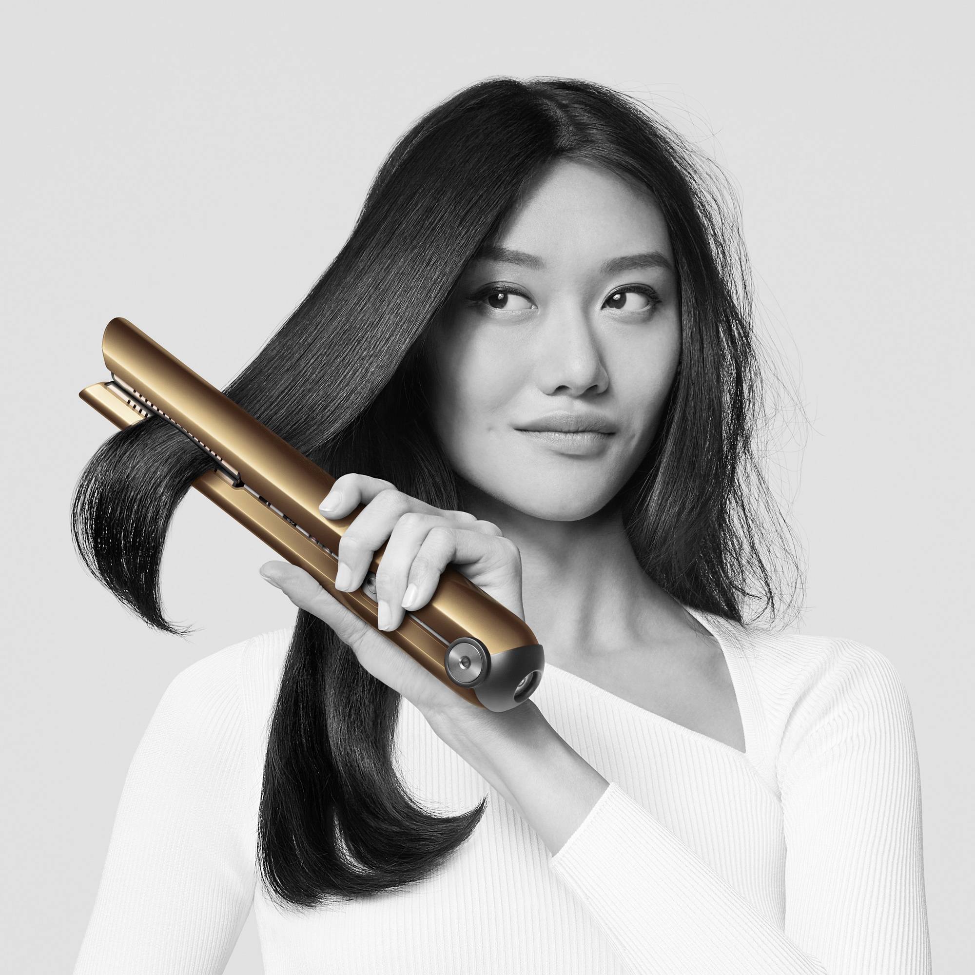 Eine Frau mit langen Haaren glättet ihr Haar mit einem goldenen Glätteisen. Sie trägt ein weißes Oberteil und schaut leicht zur Seite.