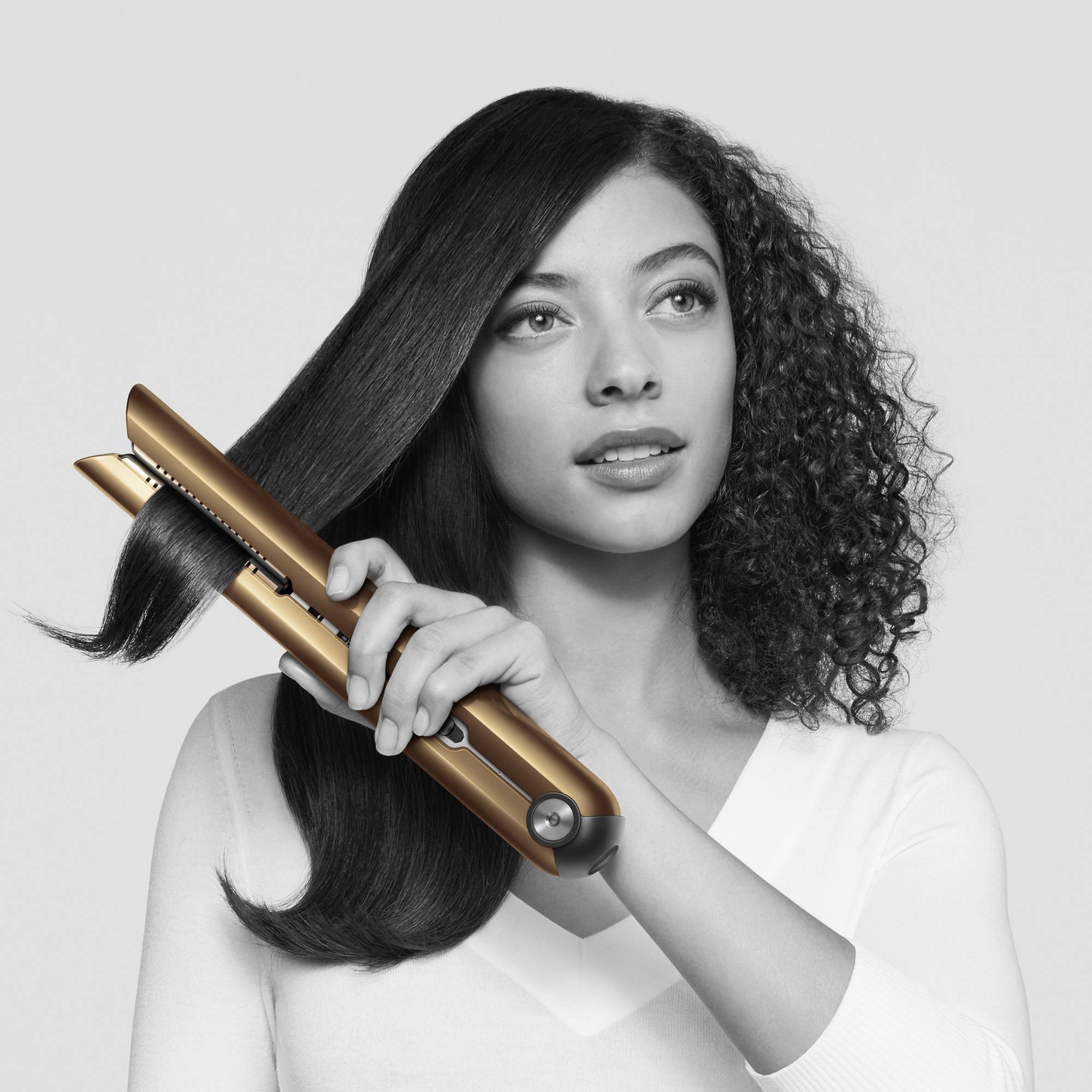 Person glättet ihr langes, welliges Haar mit einem goldenen Haarglätter. Sie trägt ein helles Oberteil und hat einen konzentrierten Ausdruck.