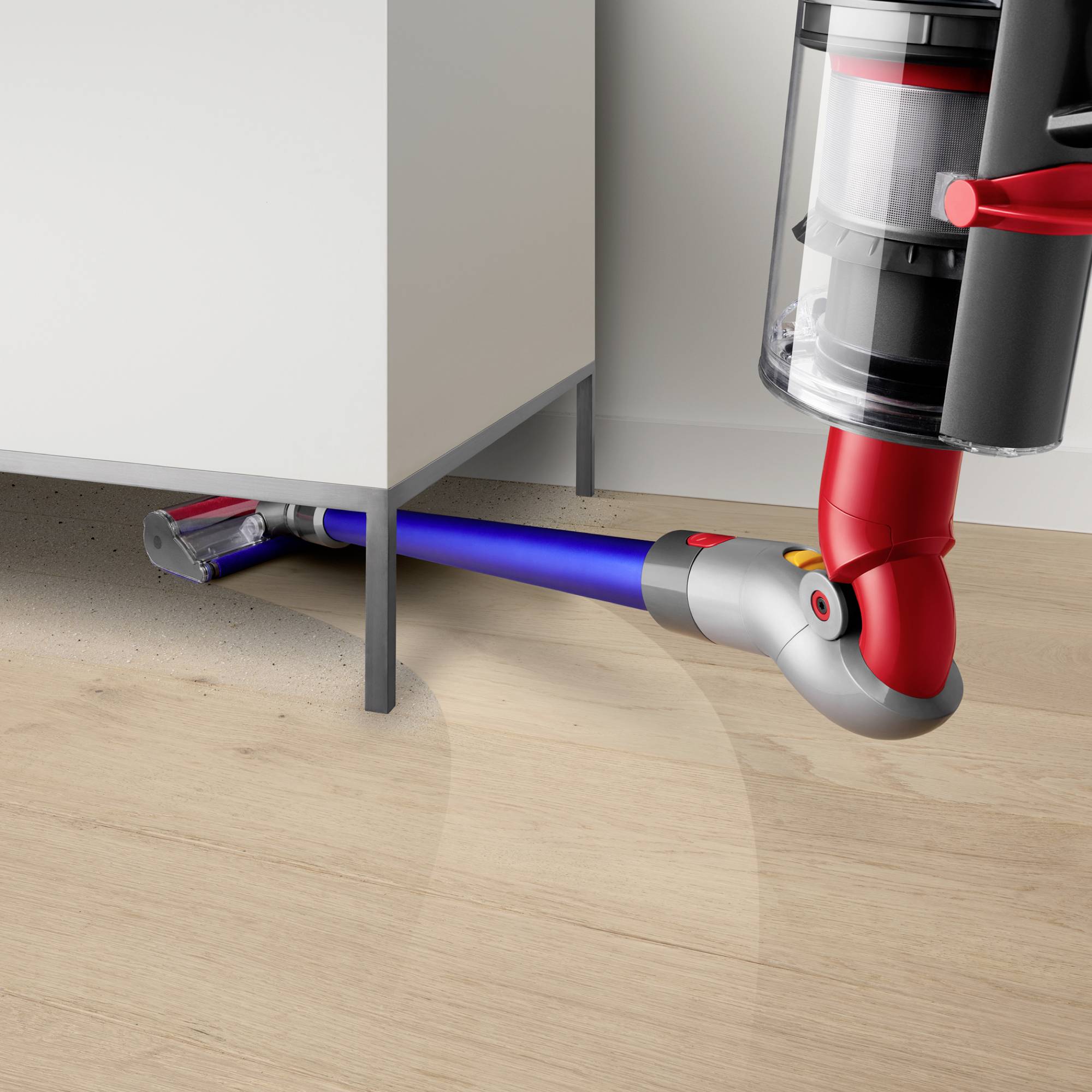 Ein kabelloser Staubsauger saugt Staub unter einem weißen Schrank auf hellem Holzboden ein. Das Gerät hat einen transparenten Behälter.
