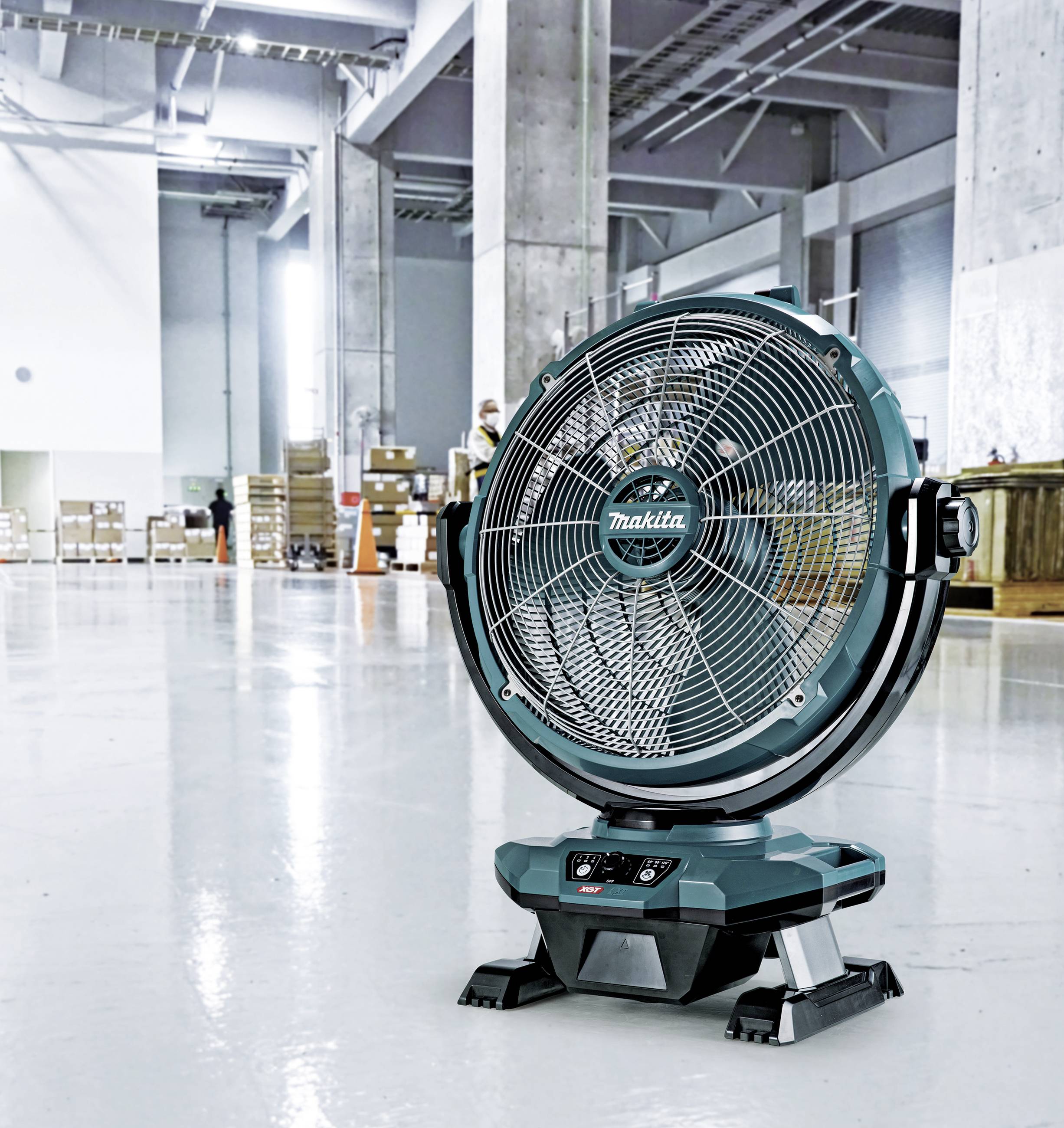 Ein grüner Industrielüfter steht auf einem glänzenden Fabrikboden mit Regalen und Kartons im Hintergrund, beleuchtet von Oberlichtern.