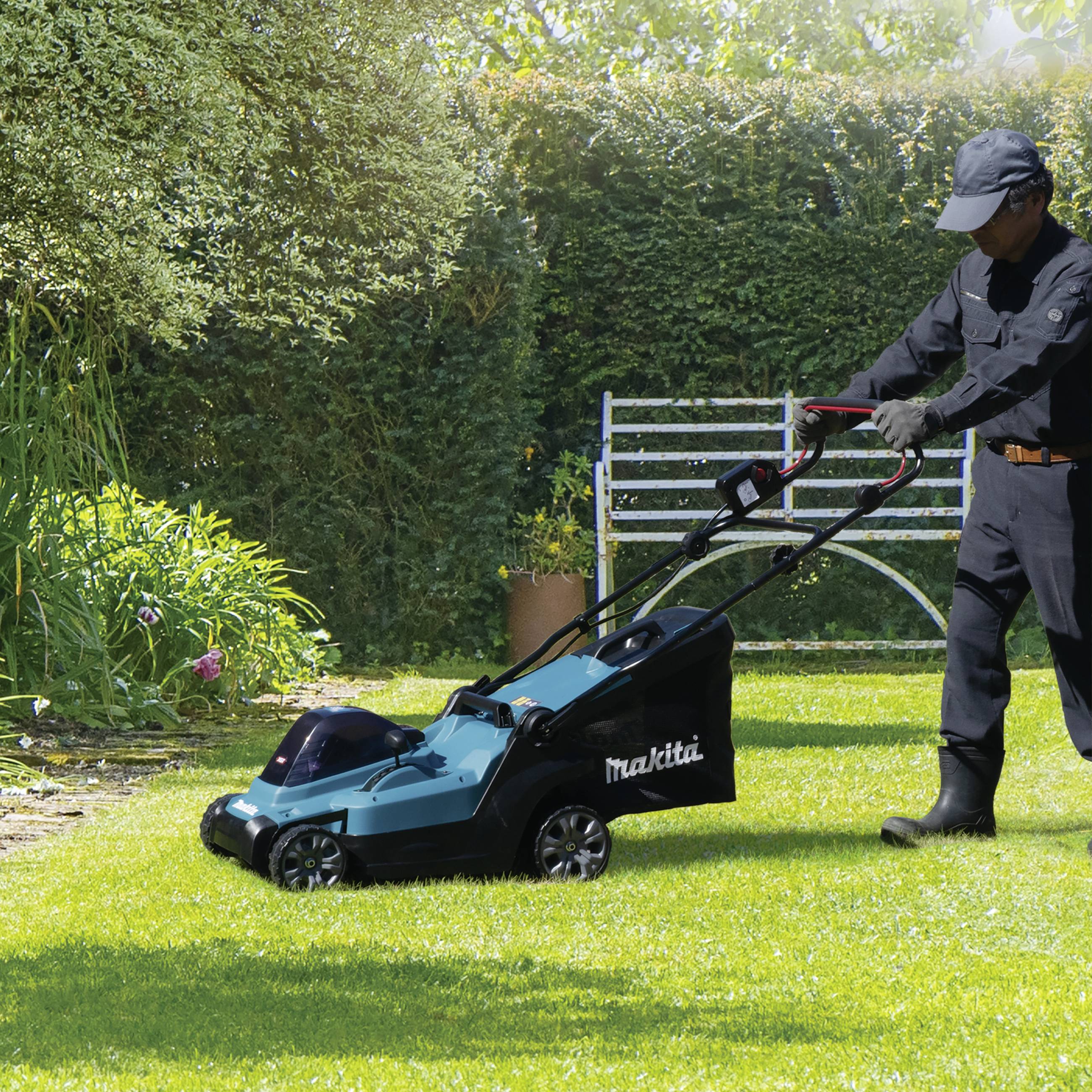 Person mäht Rasen mit elektrischem Rasenmäher auf sonnigem Gartenrasen, umgeben von grünen Pflanzen und Bäumen.
