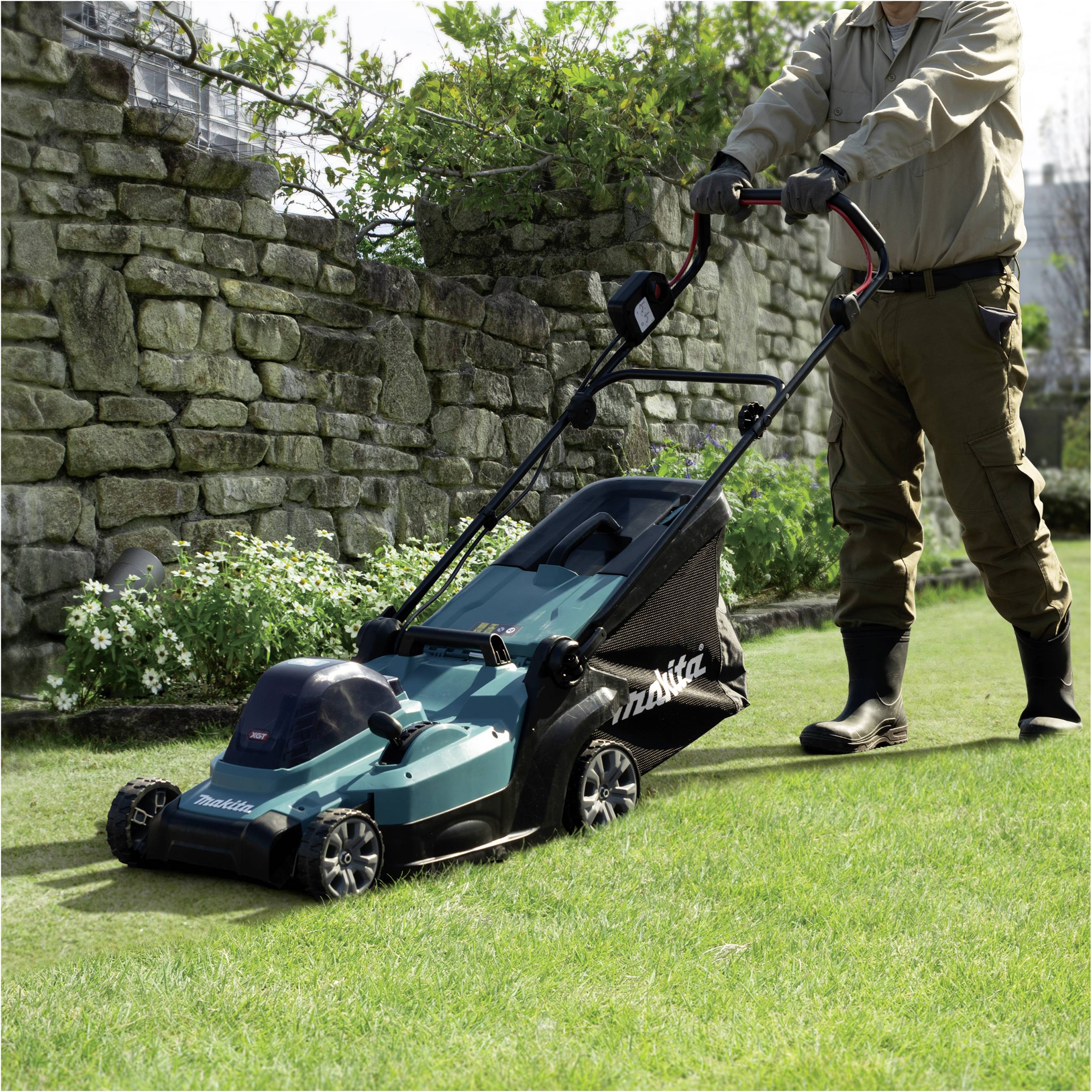Eine Person mäht mit einem elektrischen Rasenmäher das Gras vor einer Steinmauer. Der Mäher ist grün und hat einen Auffangbehälter.