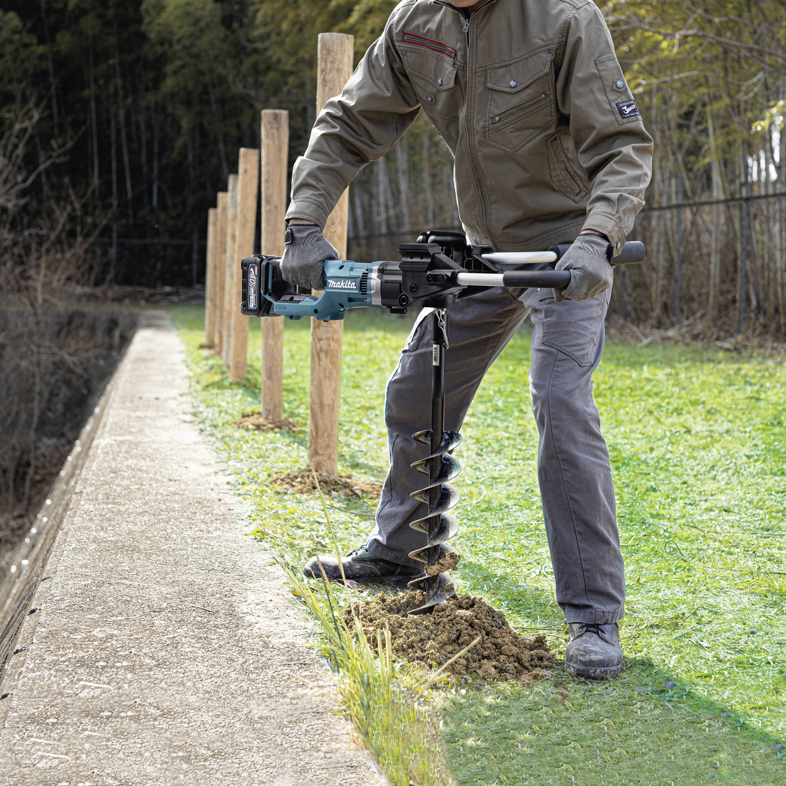 Eine Person bohrt mit einem blauen Bohrer Löcher in den Boden entlang eines Weges, im Hintergrund sind Bäume zu sehen.