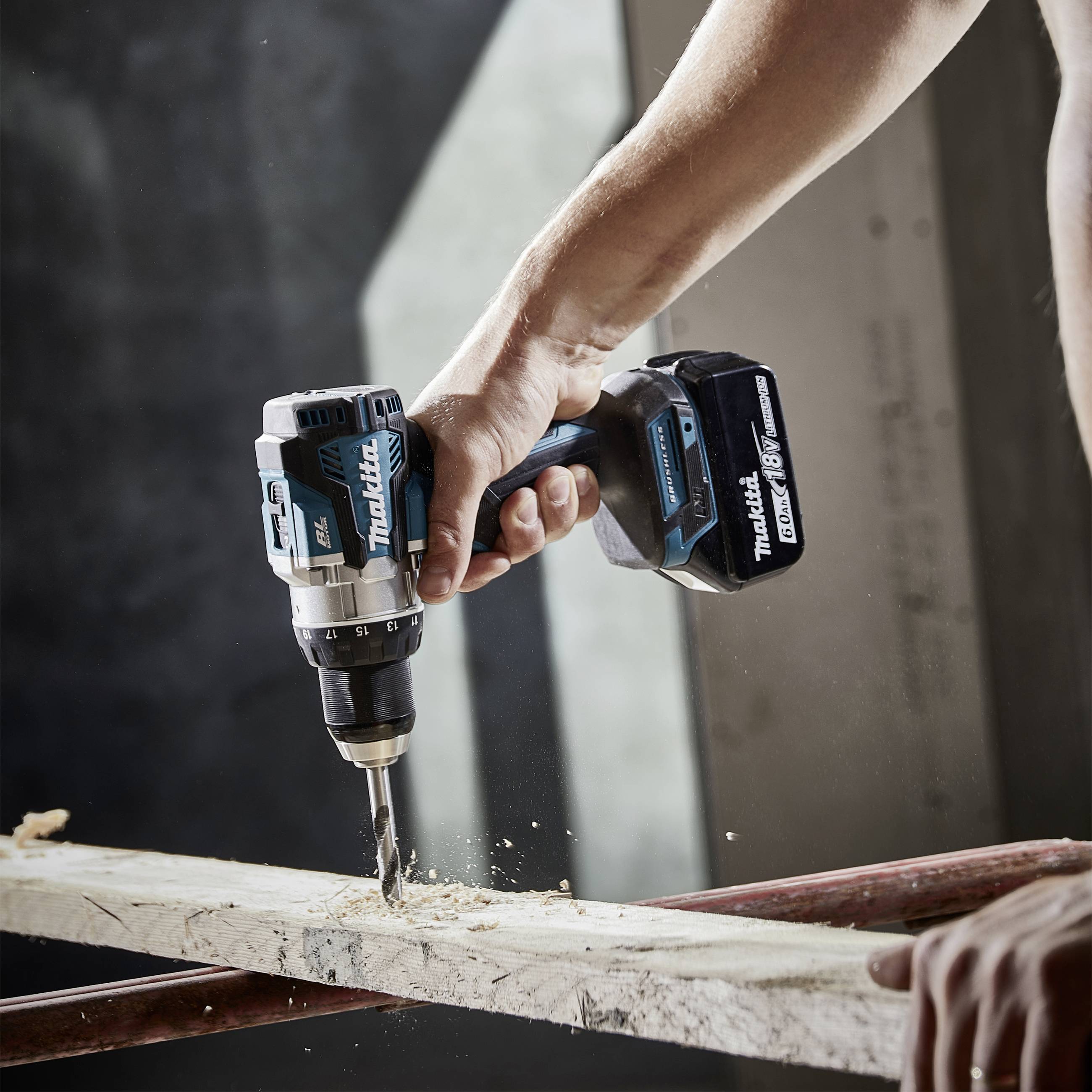 Eine Hand hält einen blauen Akkuschrauber und bohrt ein Loch in ein Holzbrett. Holzspäne fallen herab, während der Bohrer arbeitet.